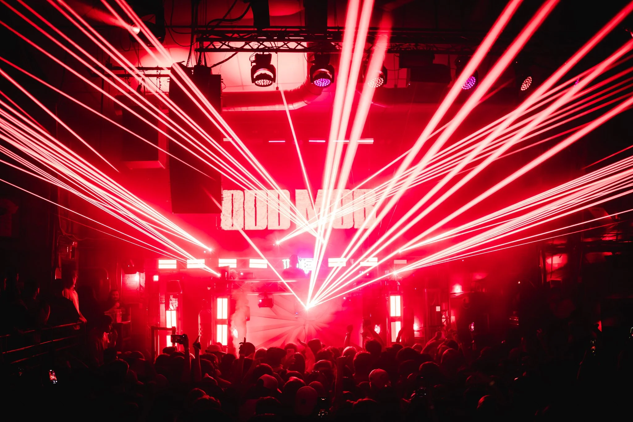 Dark nightclub with red laser lights, a crowd dancing, and a DJ on stage with a large screen displaying 'IDK' in the background.