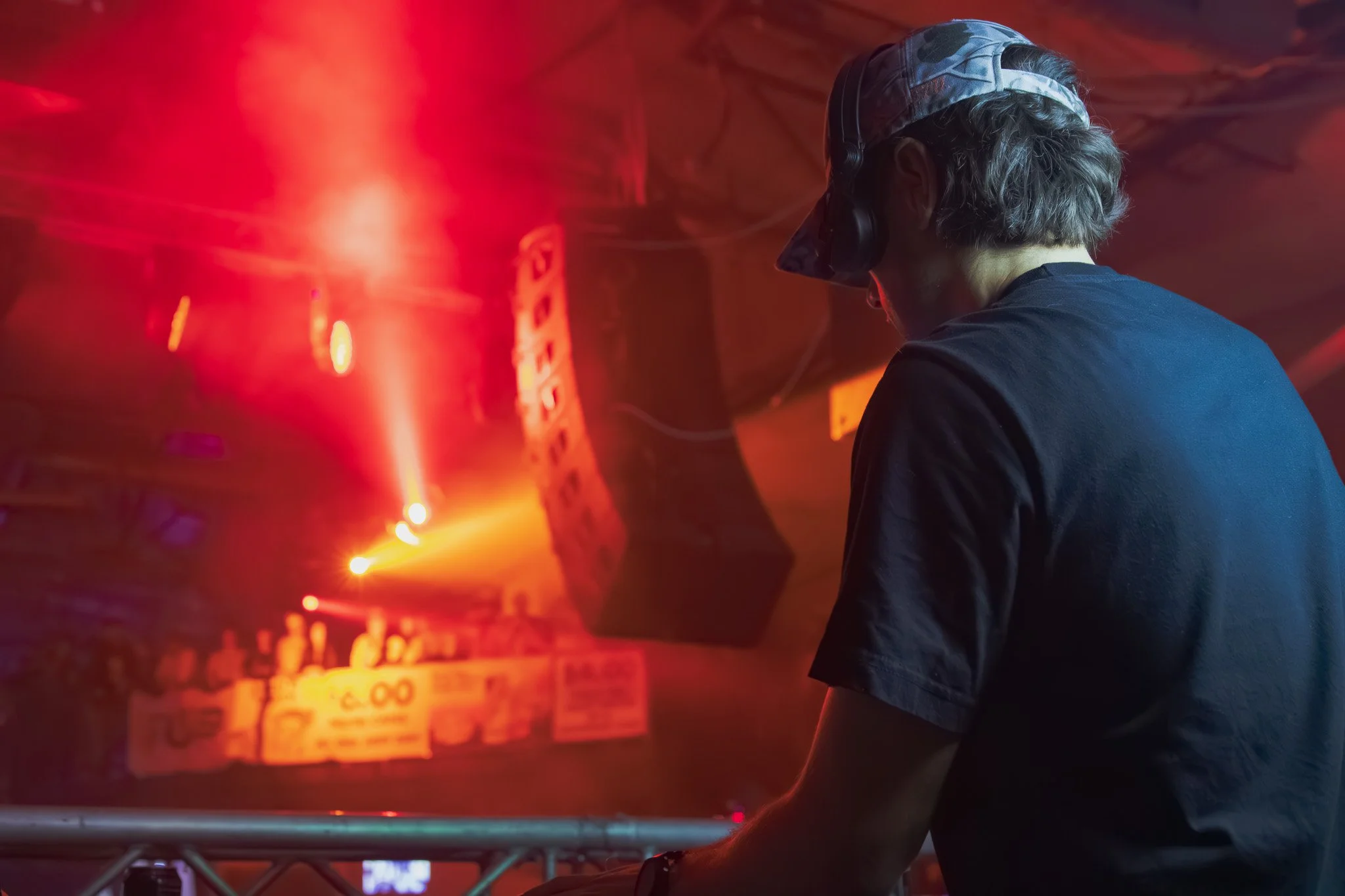 A DJ wearing headphones and a cap performing at a nightclub with colorful red and orange lights and a crowd in the background.