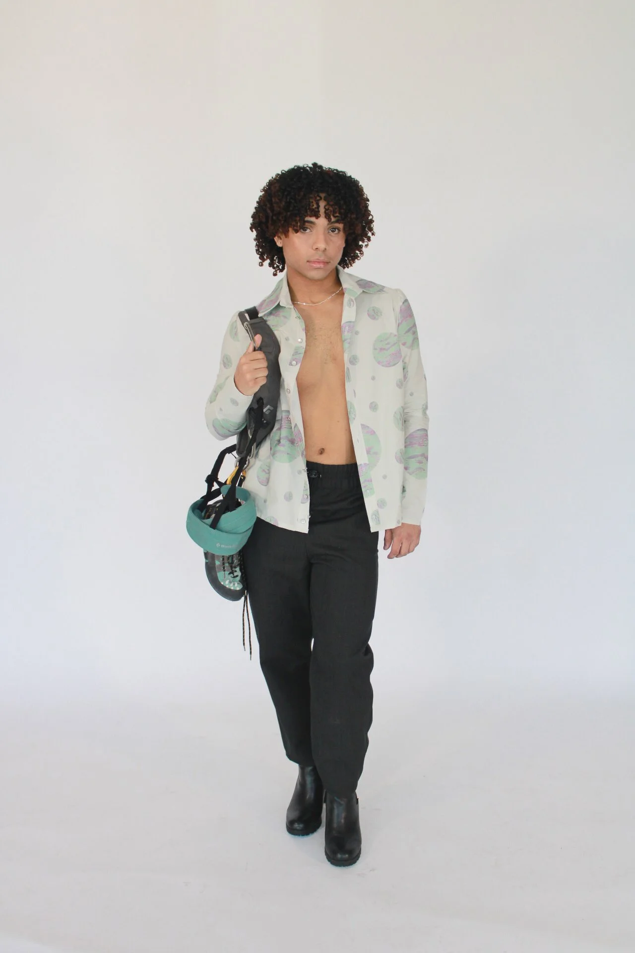 Person with curly hair, shirt open, carrying a backpack, dressed in black pants and boots, standing against a plain white background.