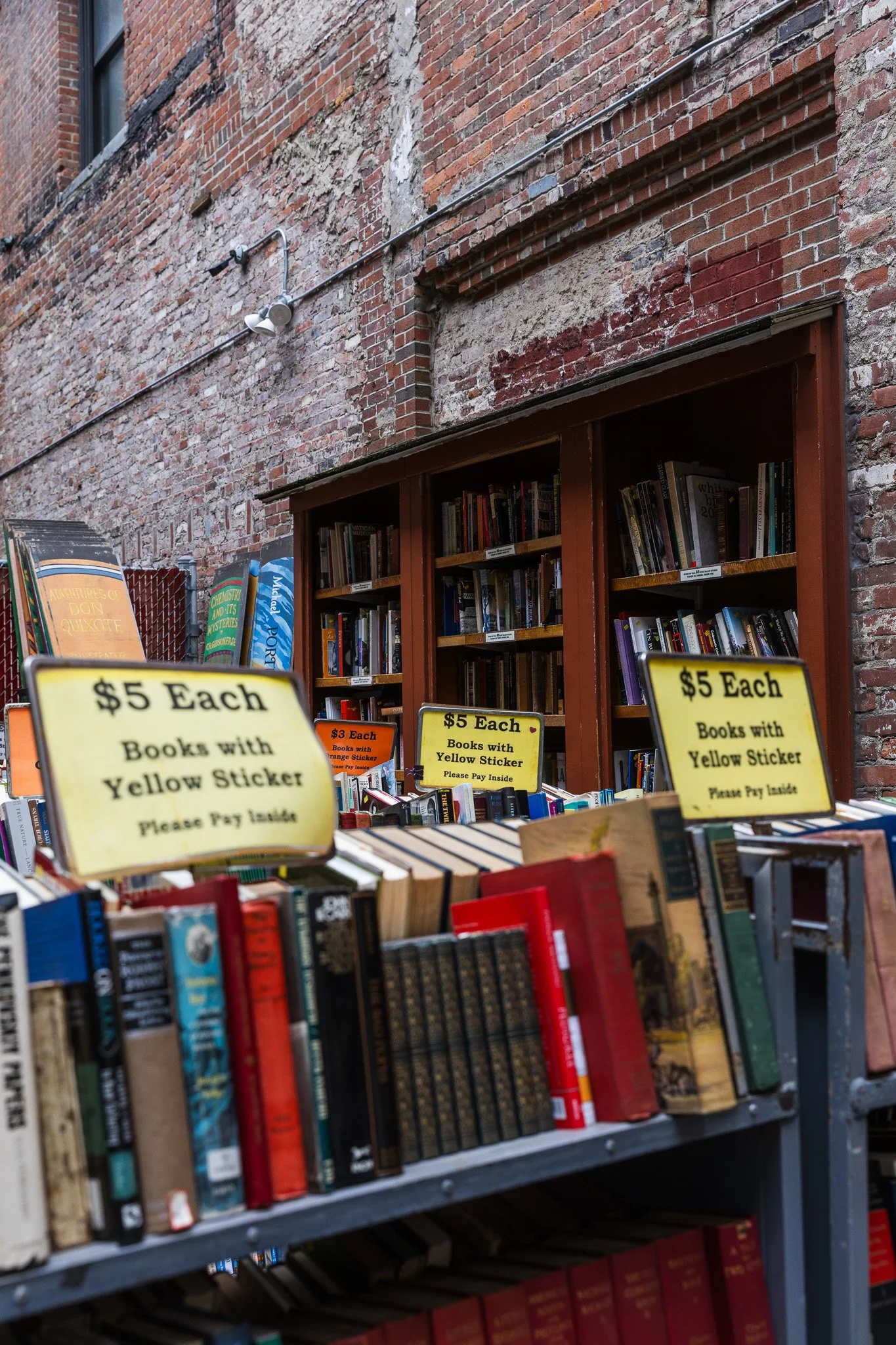 Outdoor booksale table with books priced at five dollars each, labeled with yellow stickers, set against a brick wall with a window and bookshelves.
