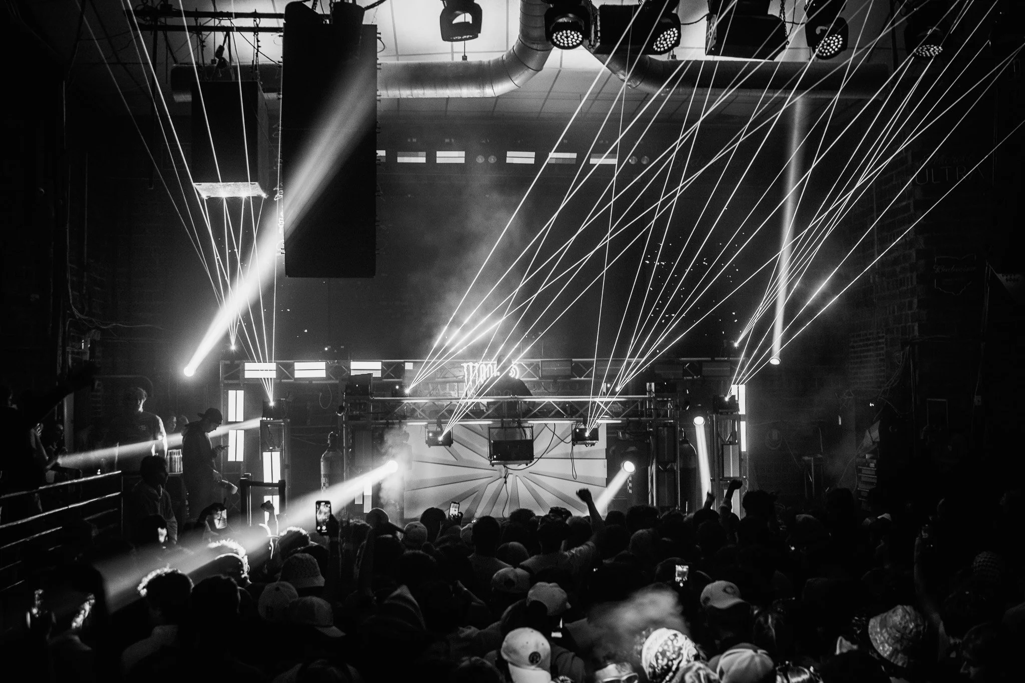 Black and white photo of a crowded indoor concert with a stage, DJ, and dynamic light effects including laser beams and spotlights.