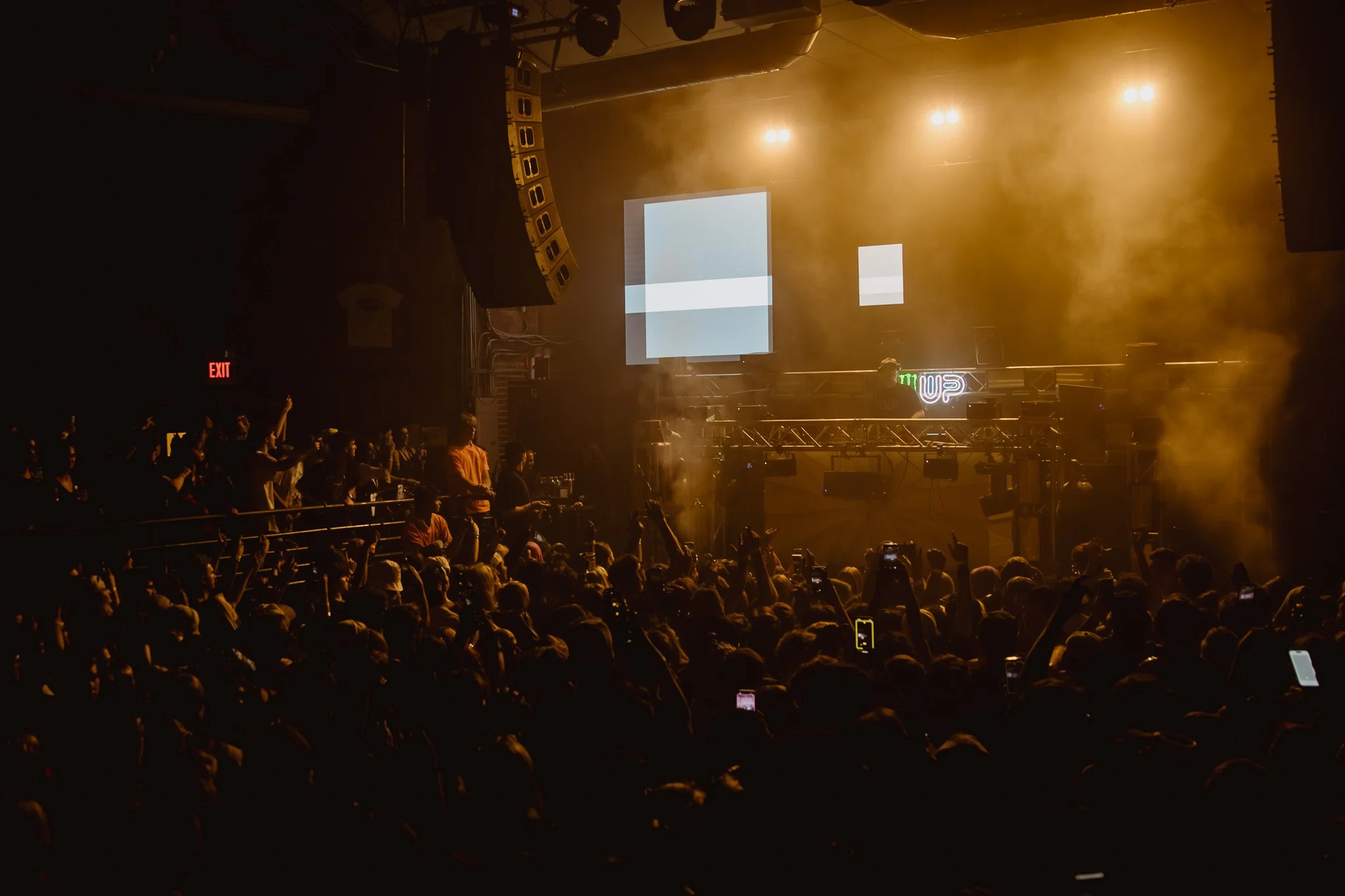 Crowd of people at a concert or music event with stage, DJ, and yellow lighting, some holding up phones.