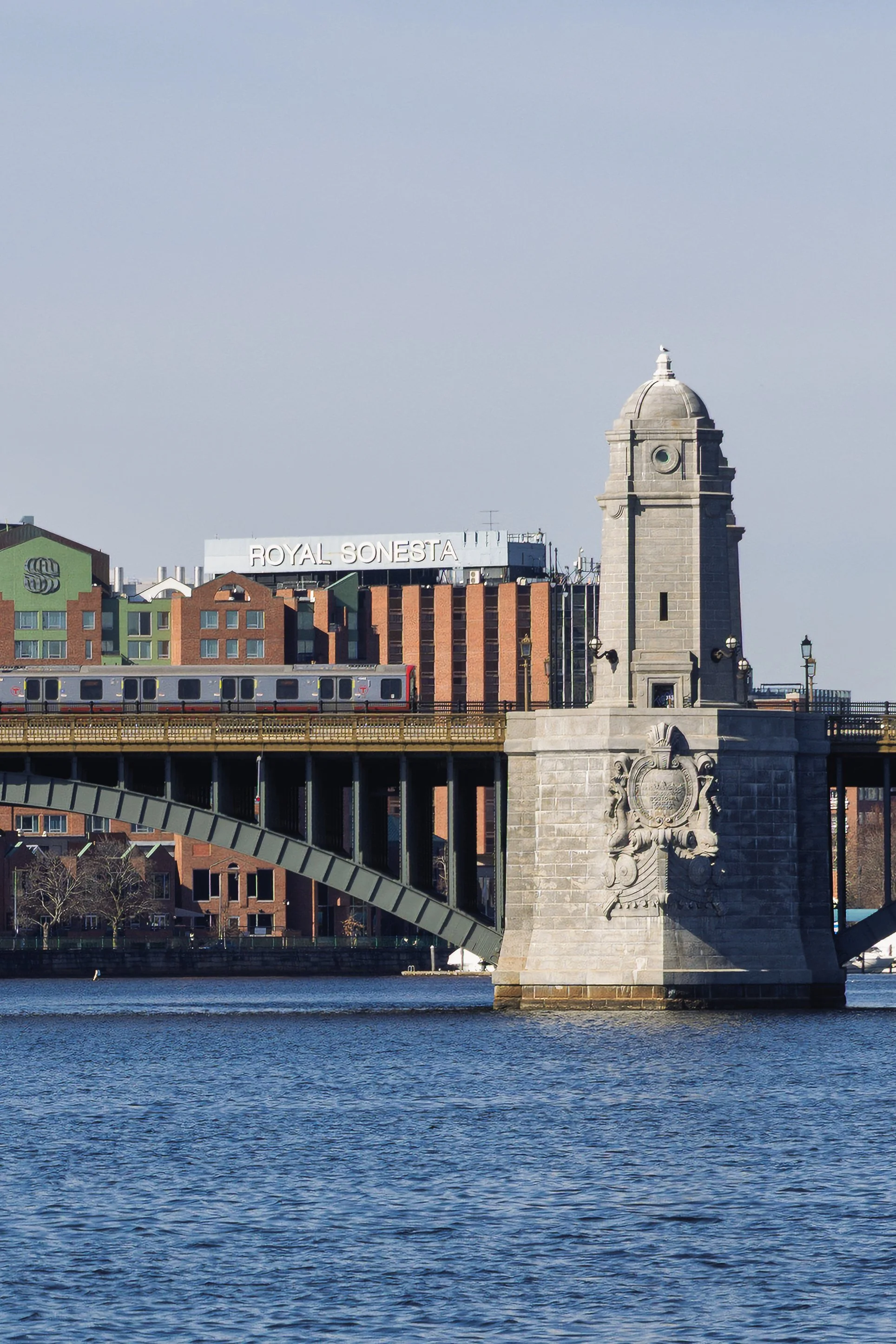 Photo of a bridge over a river with a stone tower on the right, a train crossing the bridge, and modern buildings in the background, including one with a sign that reads 'Royal Sonesta'.