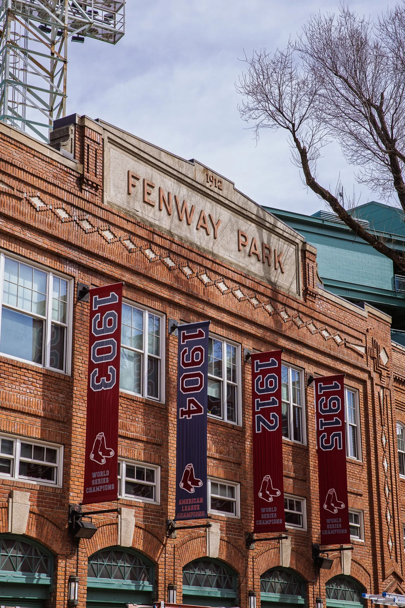 The facade of Fenway Park featuring banners celebrating the Boston Red Sox's championships in 1915, 1912, 1904, 1903, and 1915, with the park's name displayed at the top and a tree to the side.