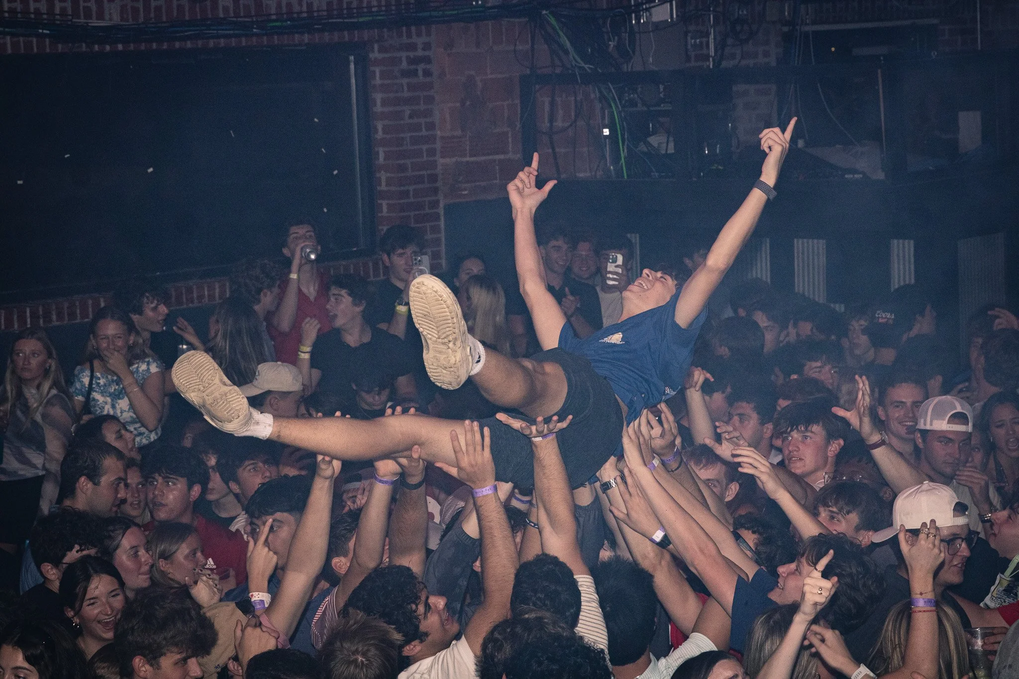 Crowd of young people at a concert or party lifting a person above their heads, who is smiling and raising arms in celebration.