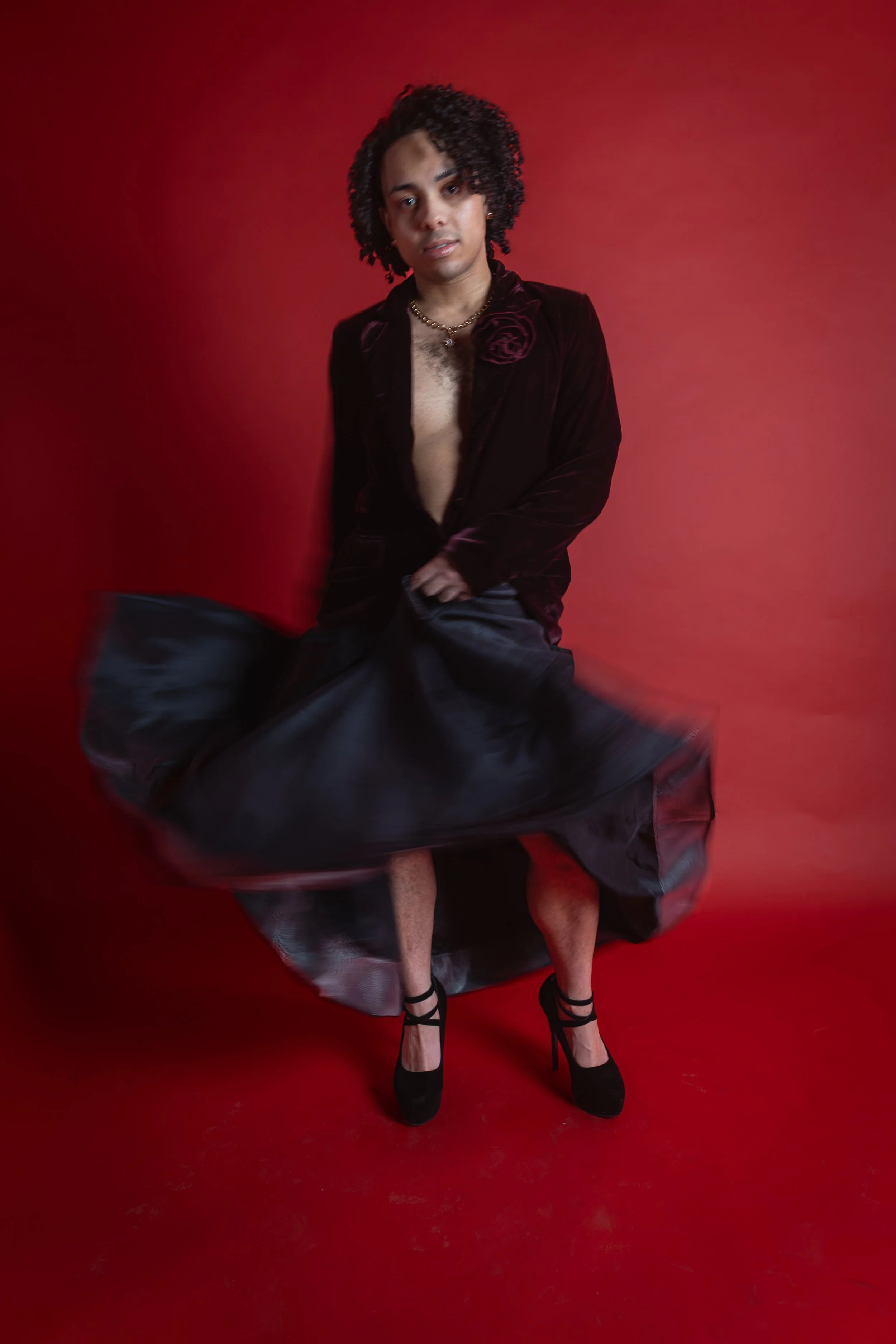 A person with curly hair, wearing a black velvet blazer with a flower detail, a gold necklace, and black high heels, standing against a red background with a flowing black skirt.