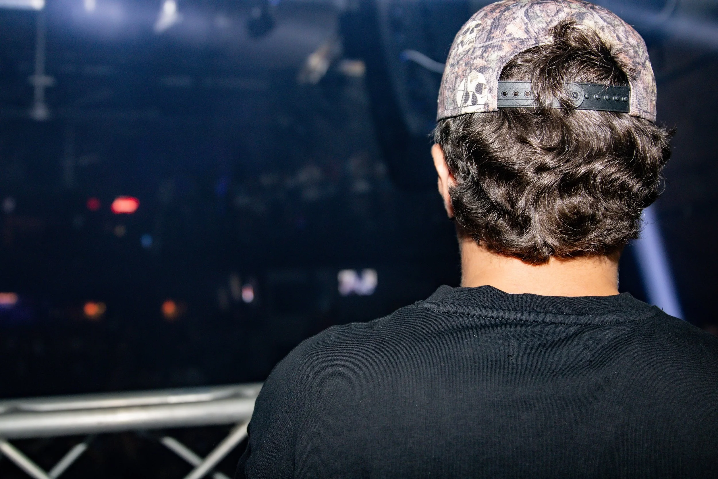 Back view of a young man wearing a camo baseball cap and a black T-shirt, sitting in front of a dark screen or monitor, with some faint lights.