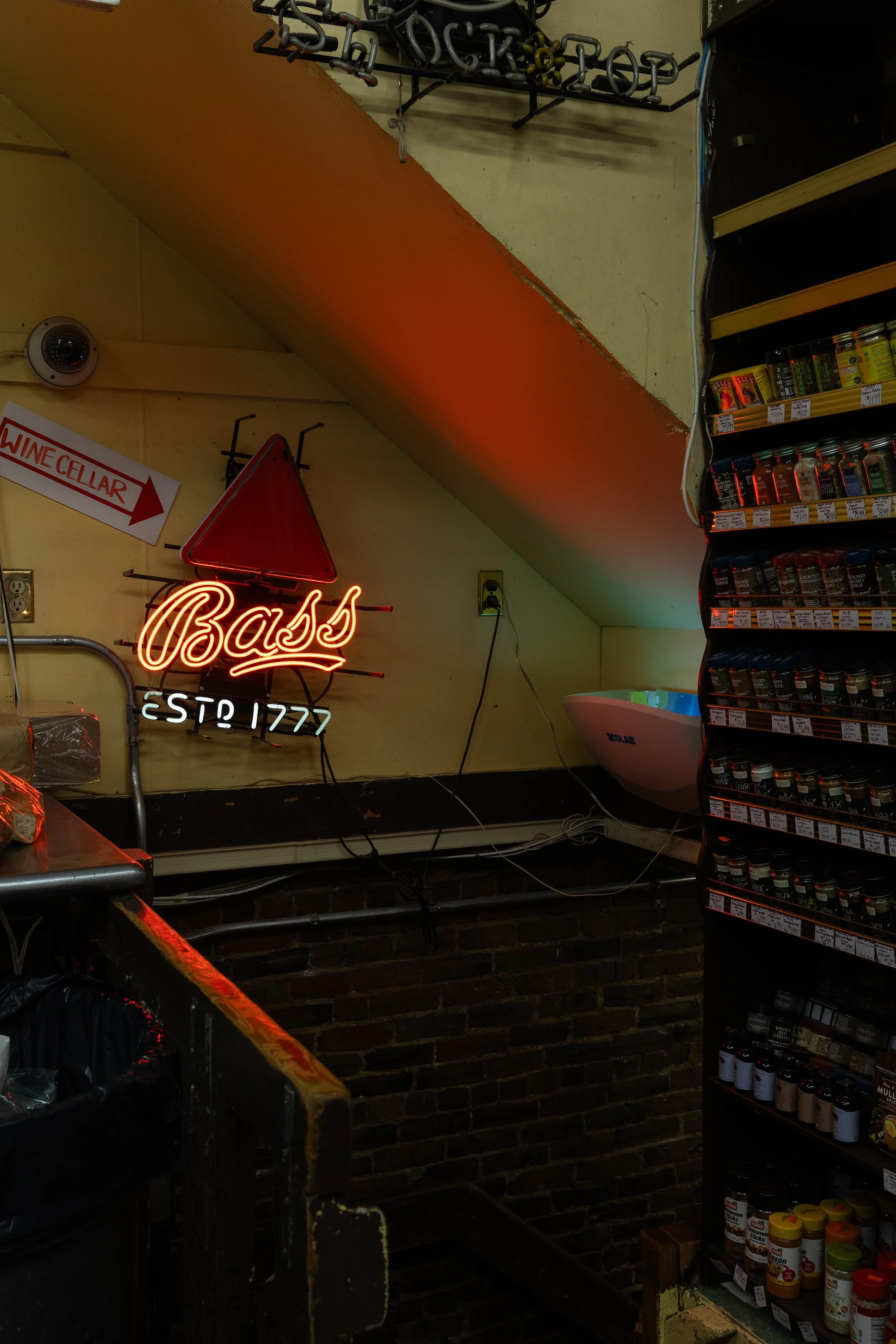 Neon sign saying 'Bass' in a store or restaurant with spice jars on the shelf to the right and a wine cellar sign pointing left.