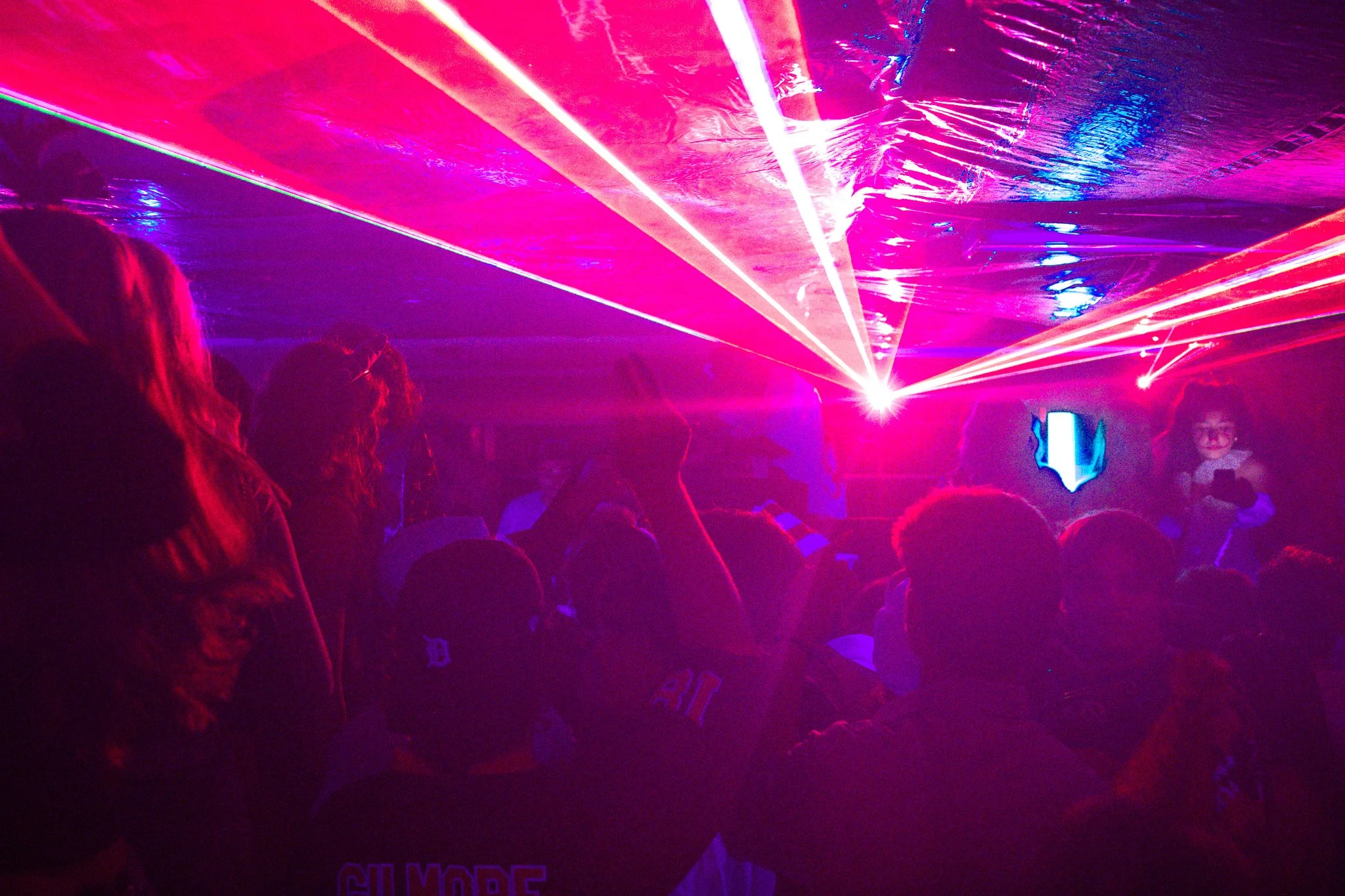 People gathered in a dark room with colorful laser lights, possibly at a party or concert, with silhouettes of attendees and laser beams in pink, purple, and blue.