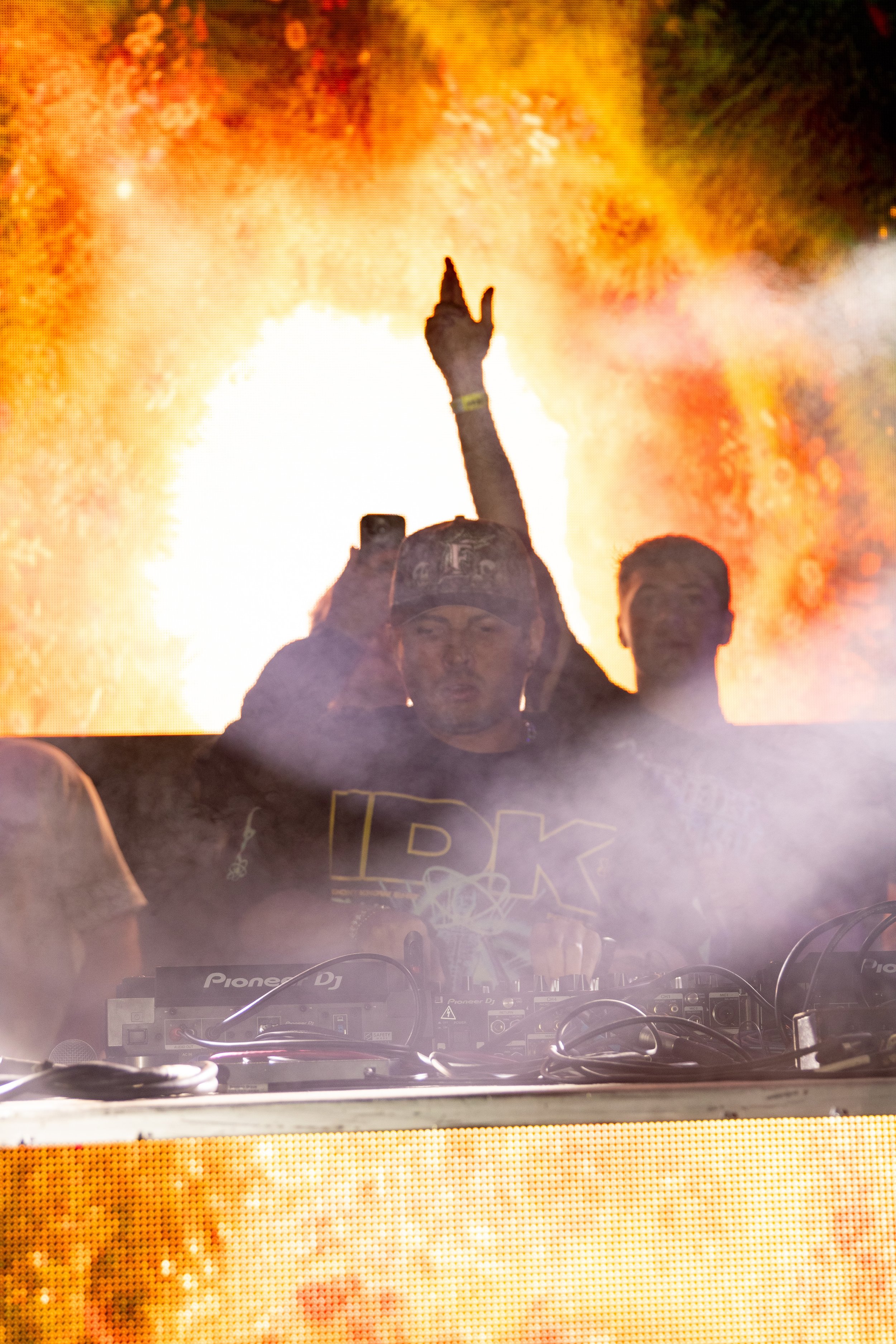 A DJ performing at a concert with a fiery, colorful background behind him. The DJ is wearing a hat and is raising one hand while focused on the equipment.