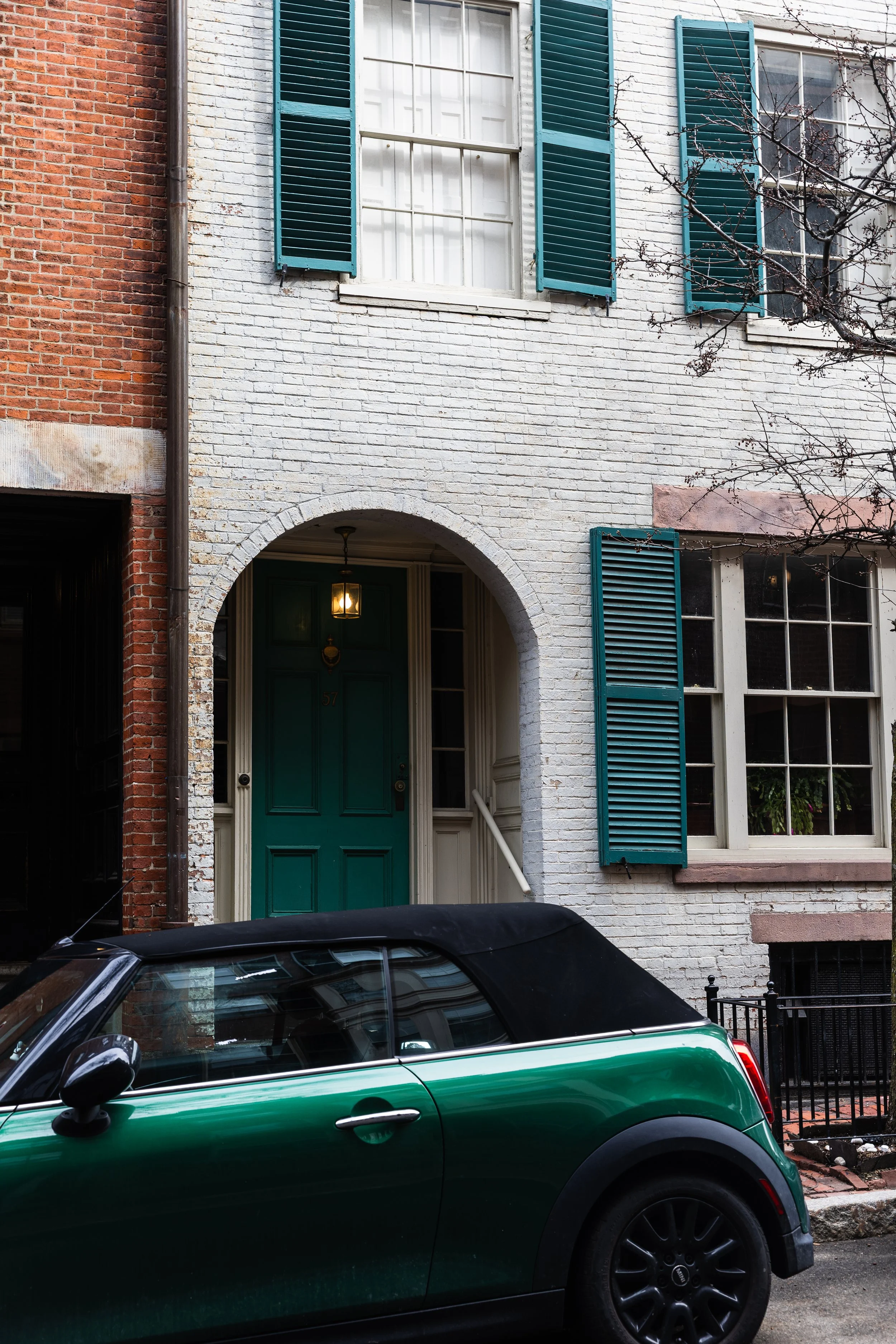 A green Mini Cooper convertible parked in front of a white brick townhouse with teal shutters and a teal front door, illuminated by a lantern-style light.