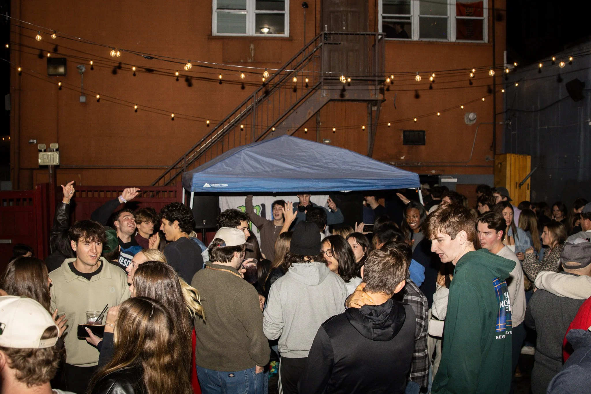 A crowded outdoor party at night with string lights hanging above and a blue canopy tent in the background. People are mingling and dancing, some holding drinks, with a building with windows and a staircase visible behind the tent.