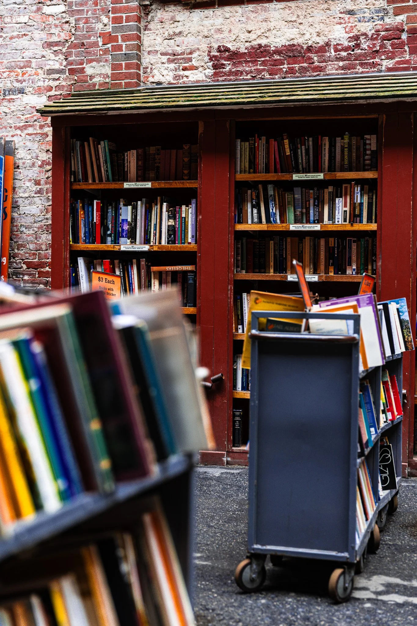 Outdoor street scene featuring a red wooden bookshelf filled with books, set against a weathered brick wall. A gray rolling cart with books is in the foreground.