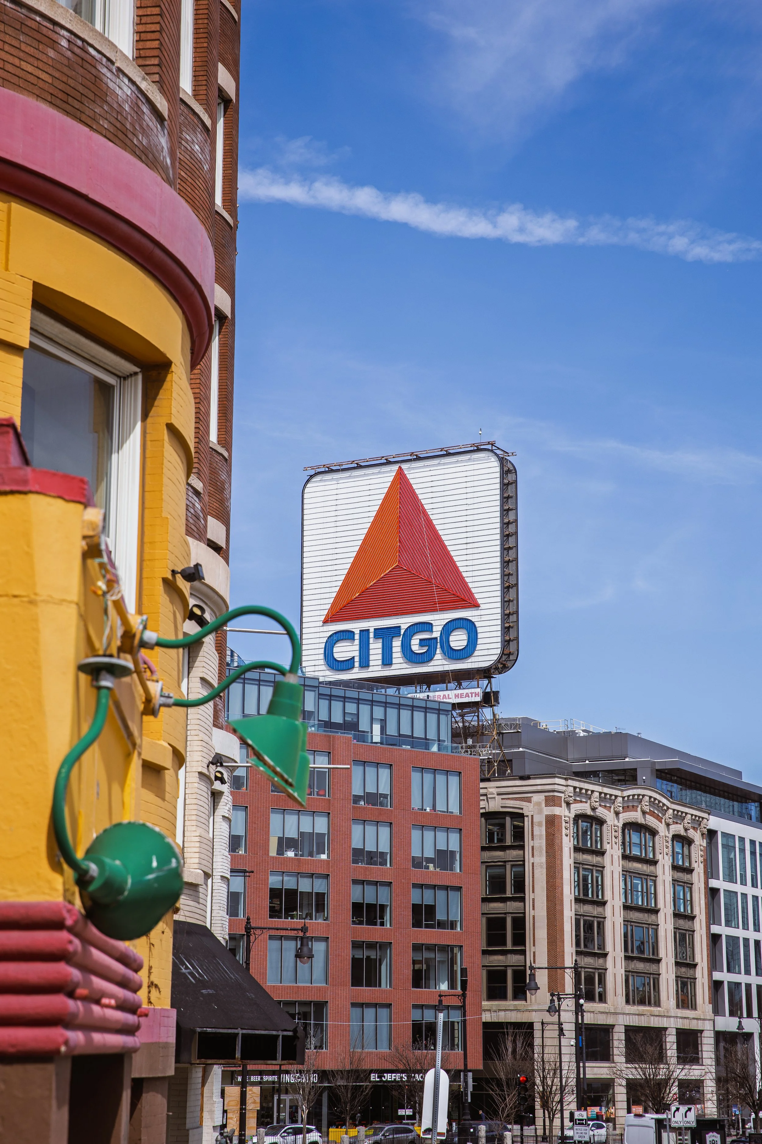 A city street with a large CITGO sign featuring a red triangular logo. Colorful buildings and a clear blue sky are in the background. Fenway Park