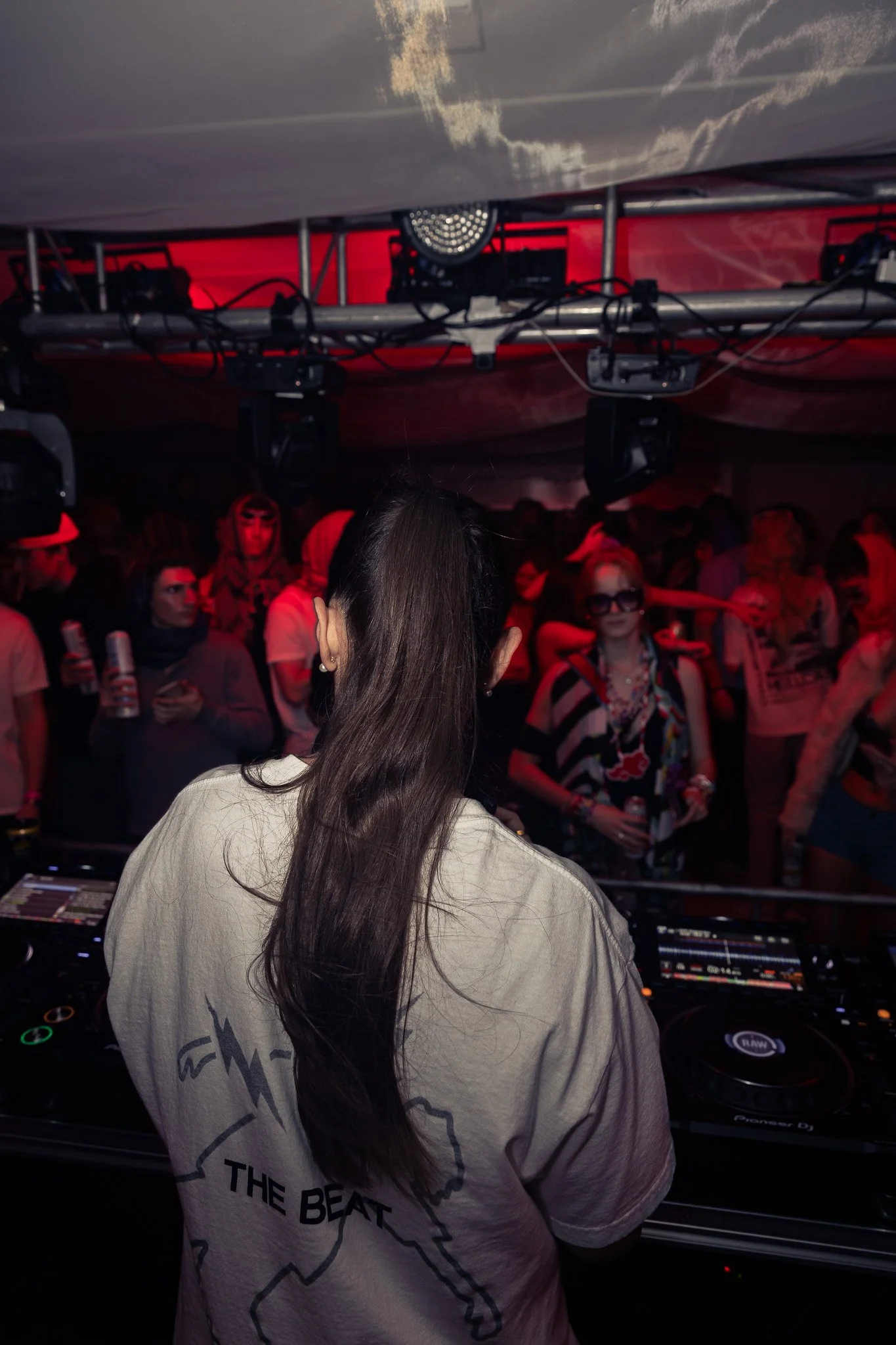 View of a DJ performing at a club with a crowd of people dancing and holding drinks under red lighting.