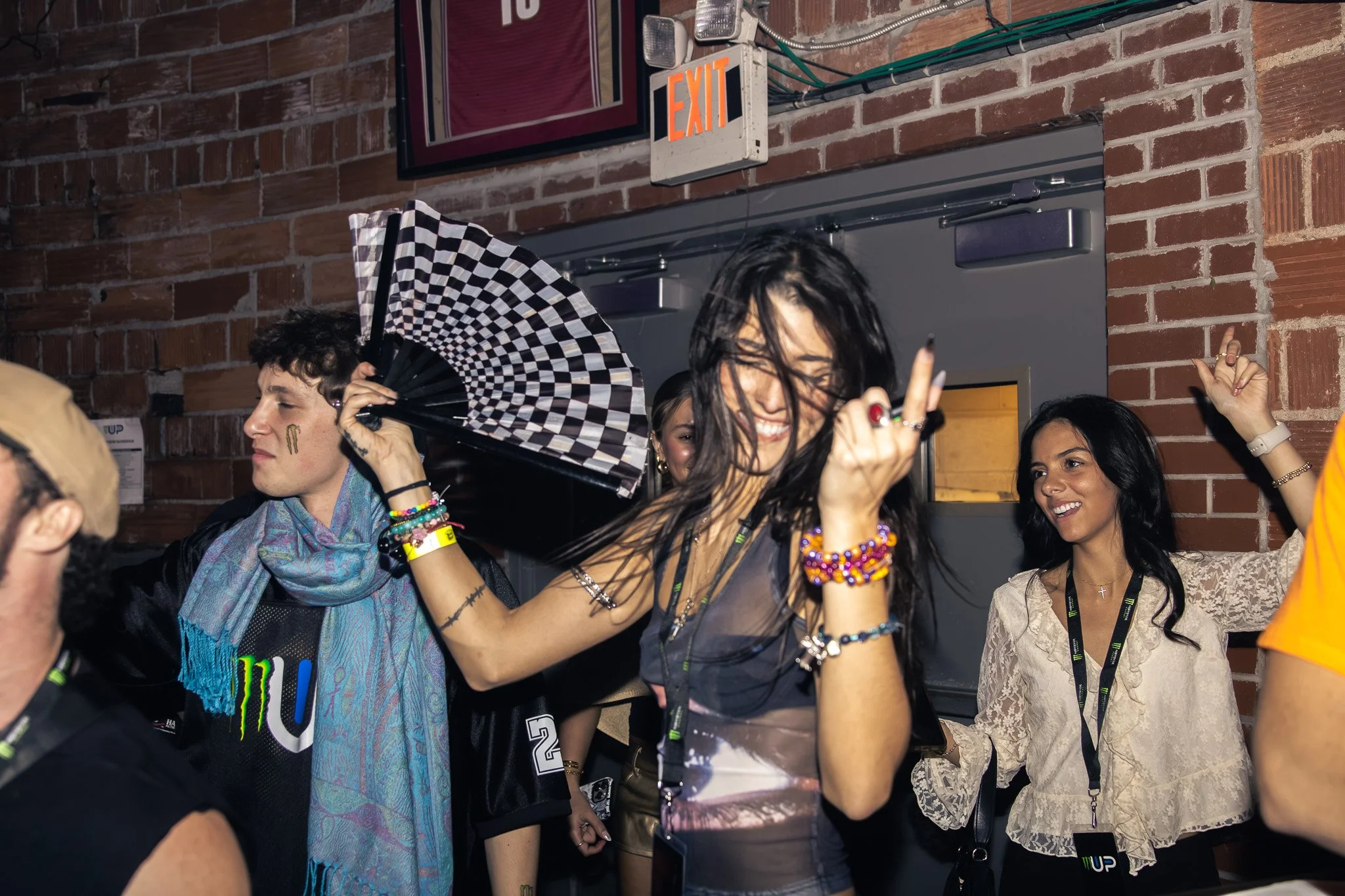 A group of young people dancing and having fun at a party or nightclub with brick walls and an exit sign in the background.