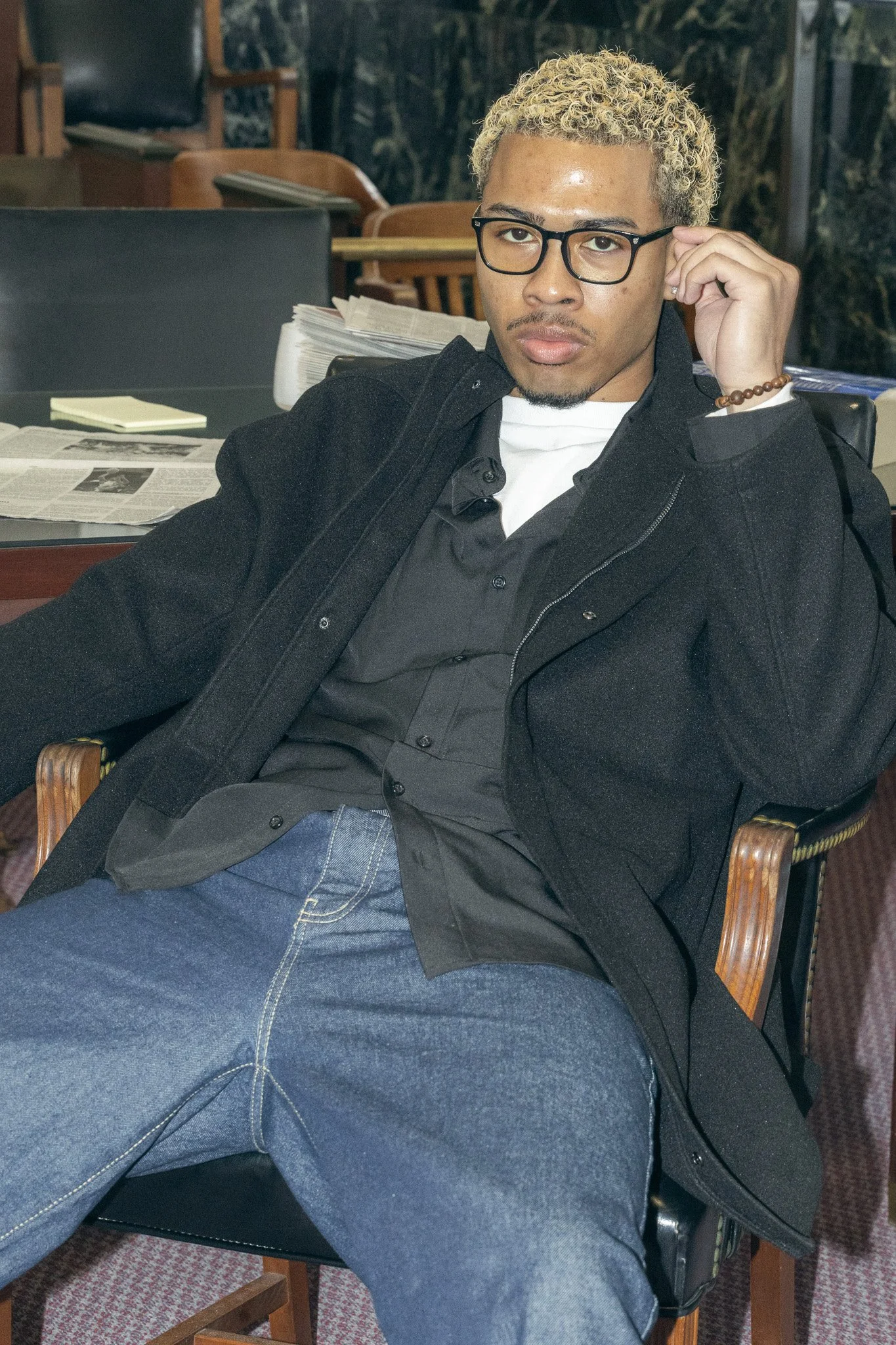 A young man with curly blonde hair, wearing glasses, a black jacket, and a black shirt, sitting in a chair in an office or library environment.