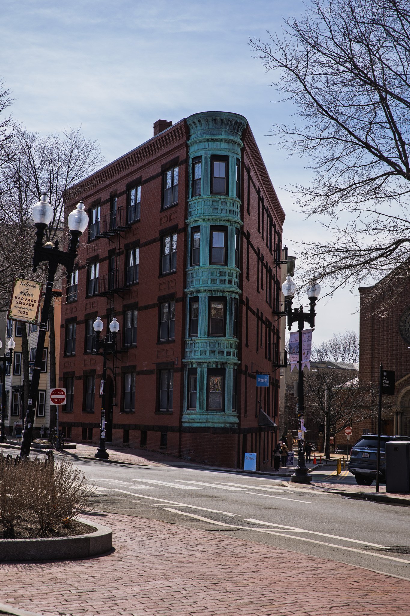 A brick building with a curved bay window painted in teal, situated on a city street corner with trees and street lamps.