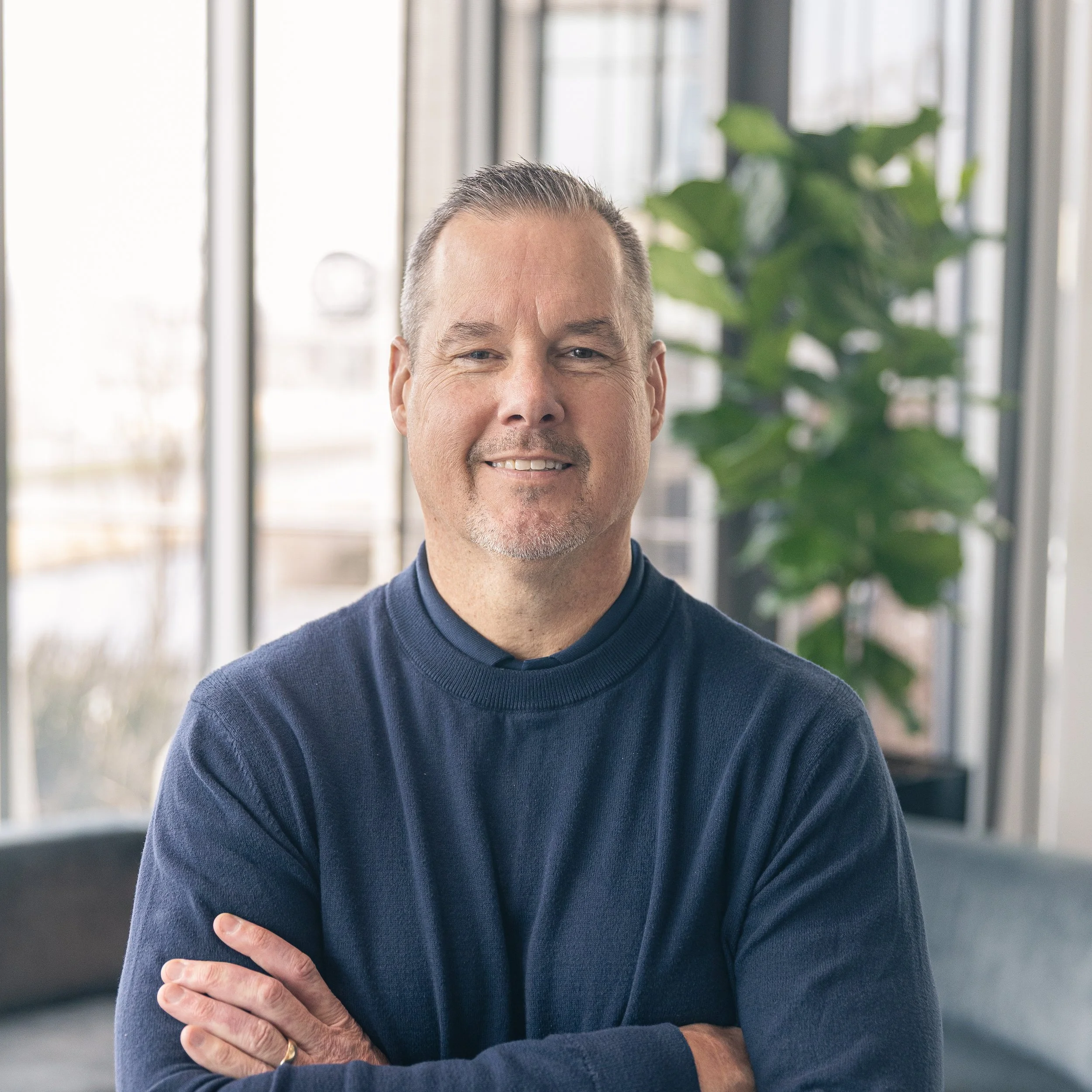 Smiling man with crossed arms in an office with large windows and a green plant in the background.