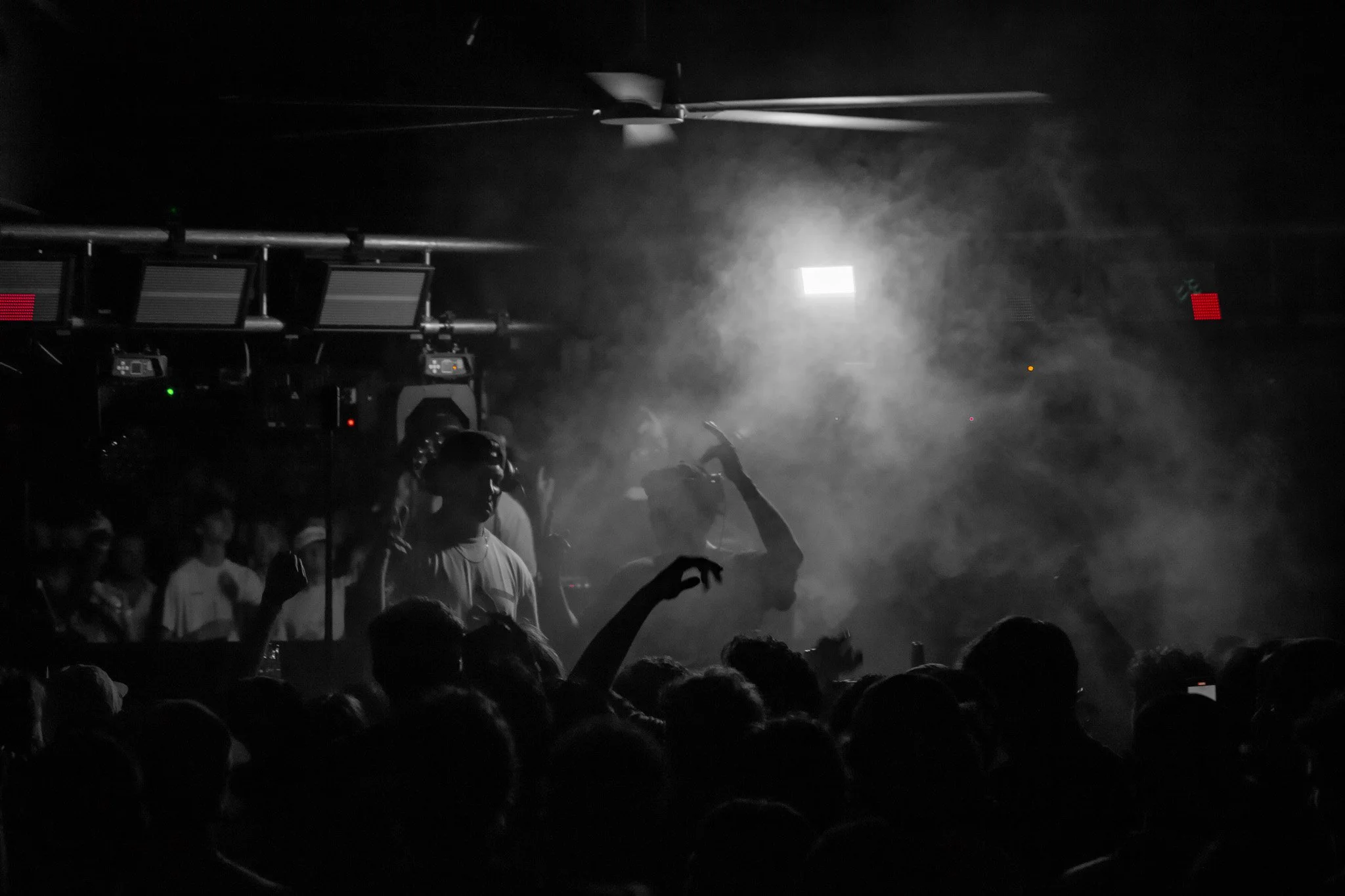 A black and white photo of a crowd at a concert or music event, with some people raising their hands, a stage or DJ booth in the background, and smoke or fog in the air.