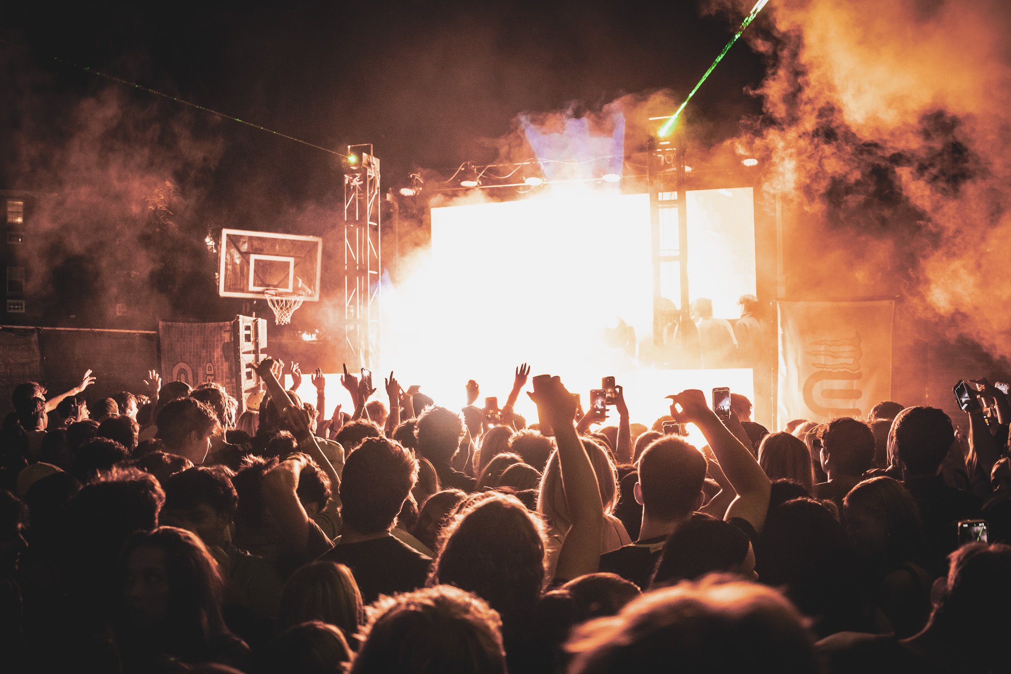 A large crowd watching a concert or music event at night with fireworks or pyrotechnics on stage, some people holding up their phones to record the performance.