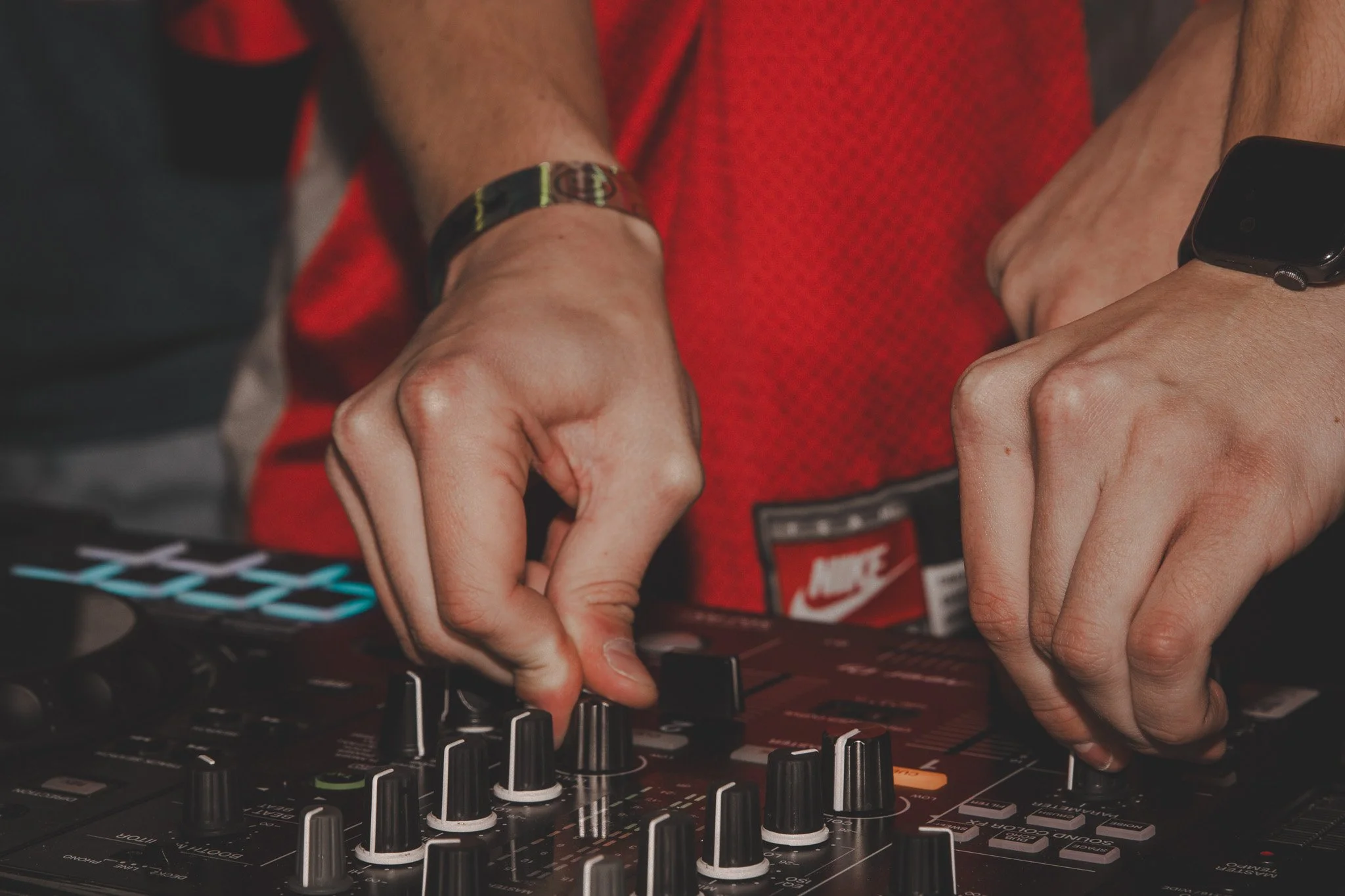 Close-up of a person adjusting controls on a DJ mixer. The individual is wearing a red shirt, a black wristband, and a smartwatch.
