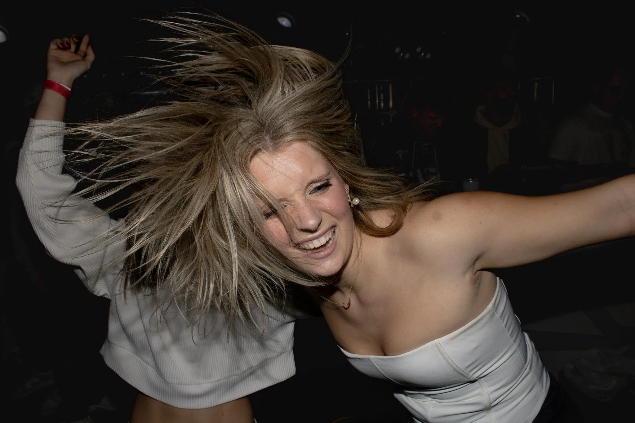 A woman with blonde hair dancing with her eyes closed and a big smile, wearing a white strapless top in a dark setting.