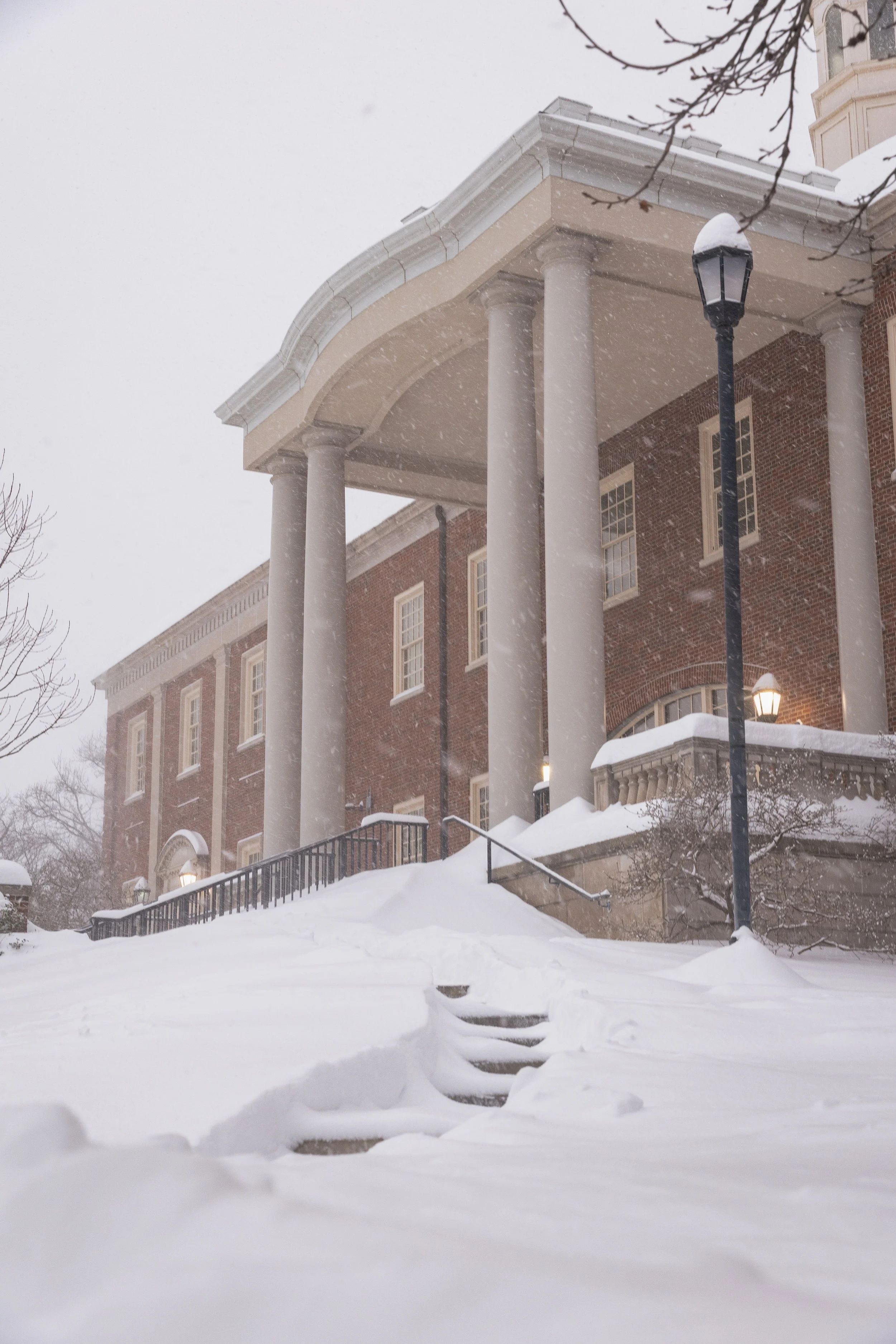 A large brick building with white columns and a portico, covered in snow during a snowy day.
