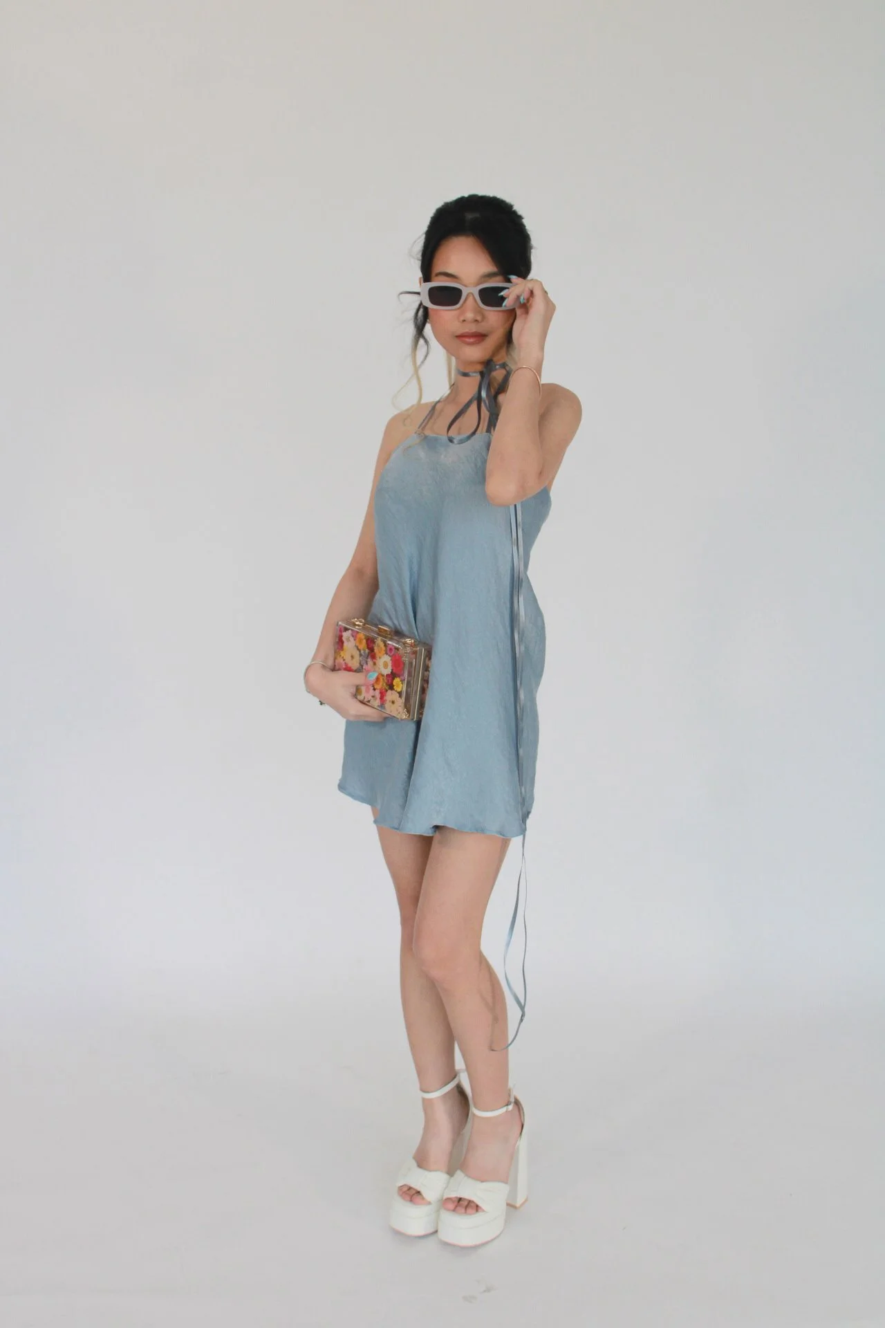 A woman in light blue dress, white platform heels, sunglasses, holding a floral clutch, posing against a plain white background.
