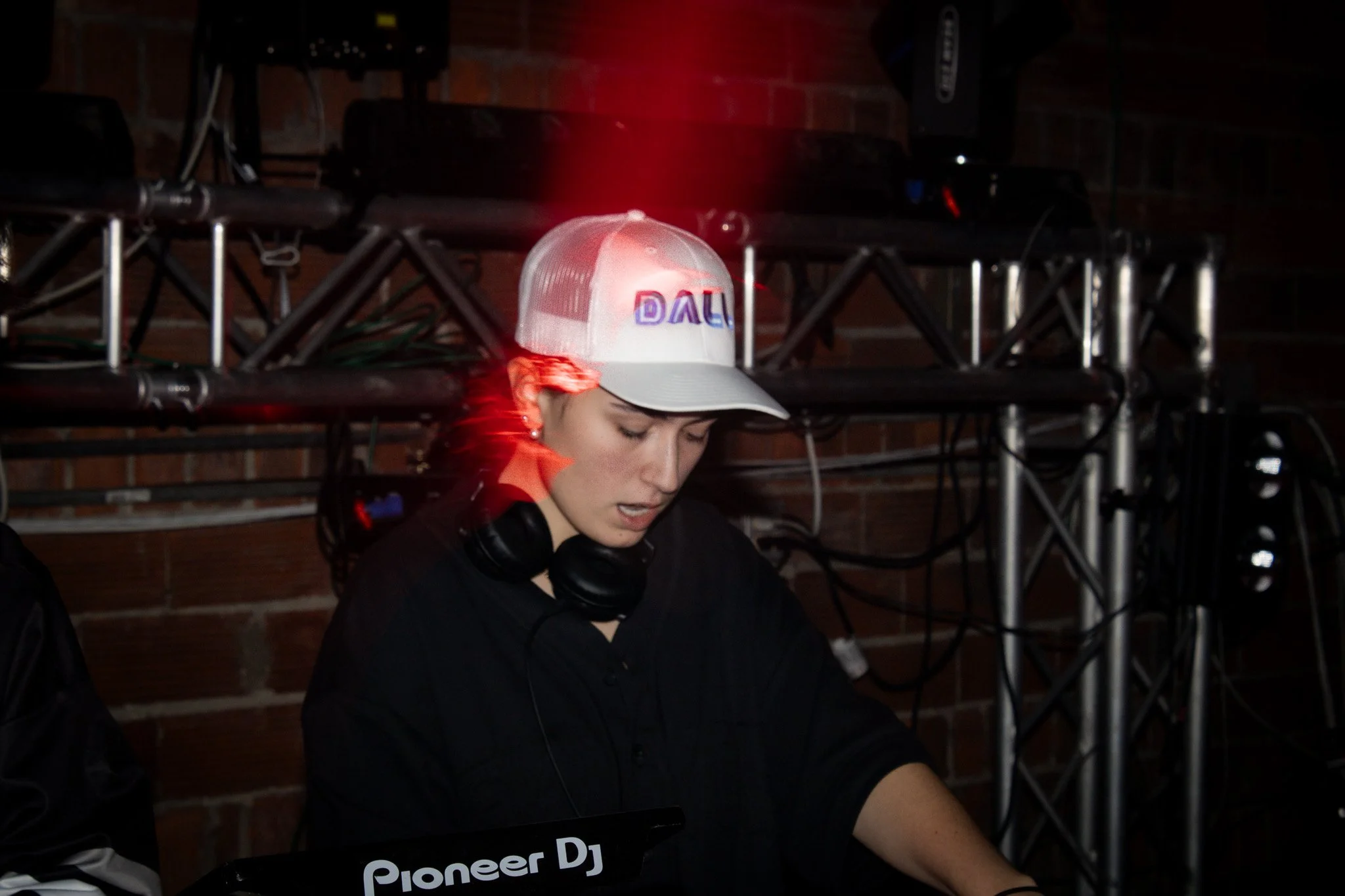 Young female DJ wearing a white trucker hat with the logo 'DALI', focused on her equipment, with headphones around her neck, in a dimly lit venue with DJ equipment and metal trusses in the background.