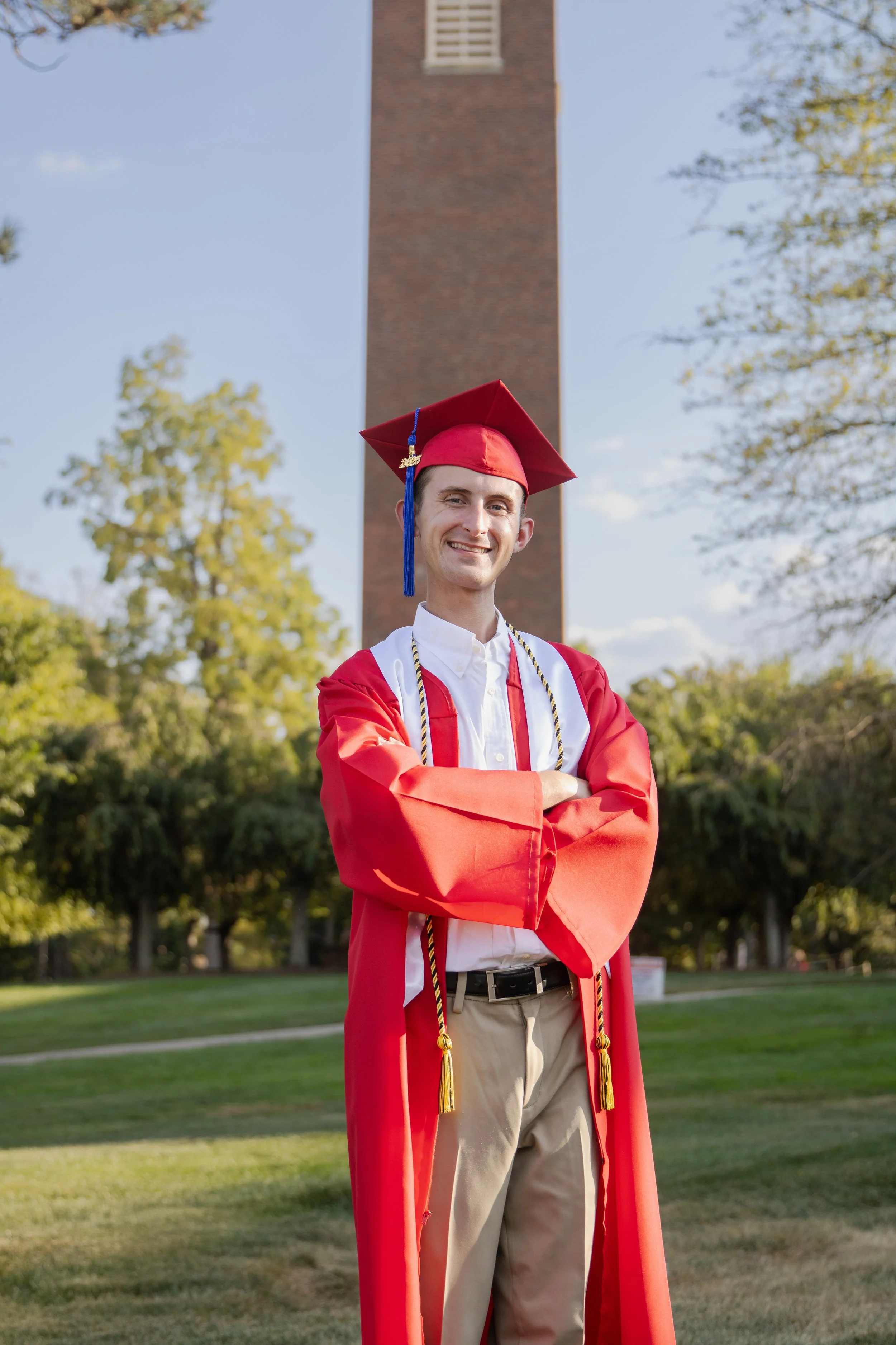 A young man in a red graduation gown and cap with a tassel, standing outdoors with arms crossed, smiling. There is a brick tower and trees with green leaves in the background.