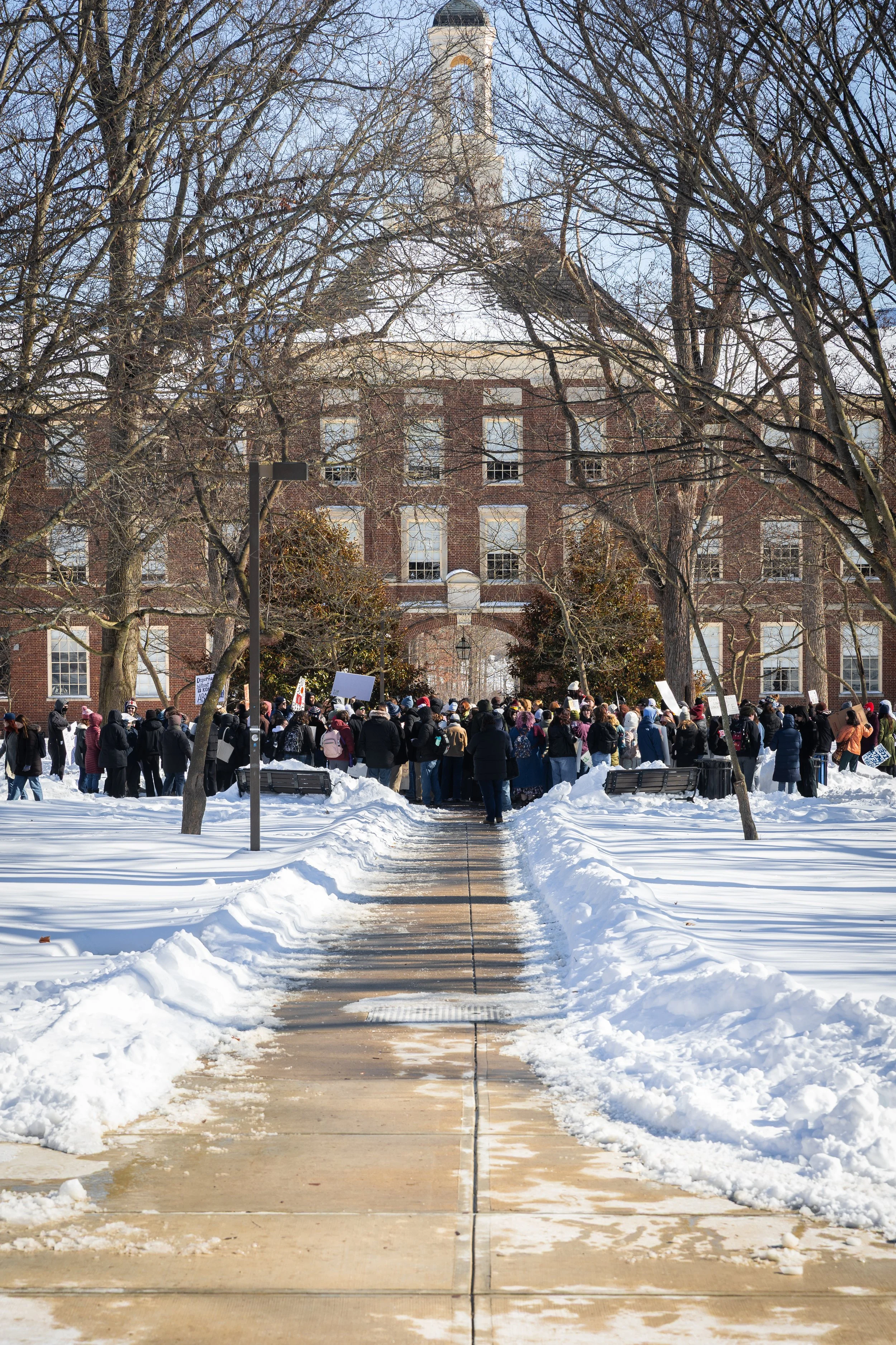 A group of people gathered outside a large brick building in winter, holding signs and protesting on a snow-covered walkway.