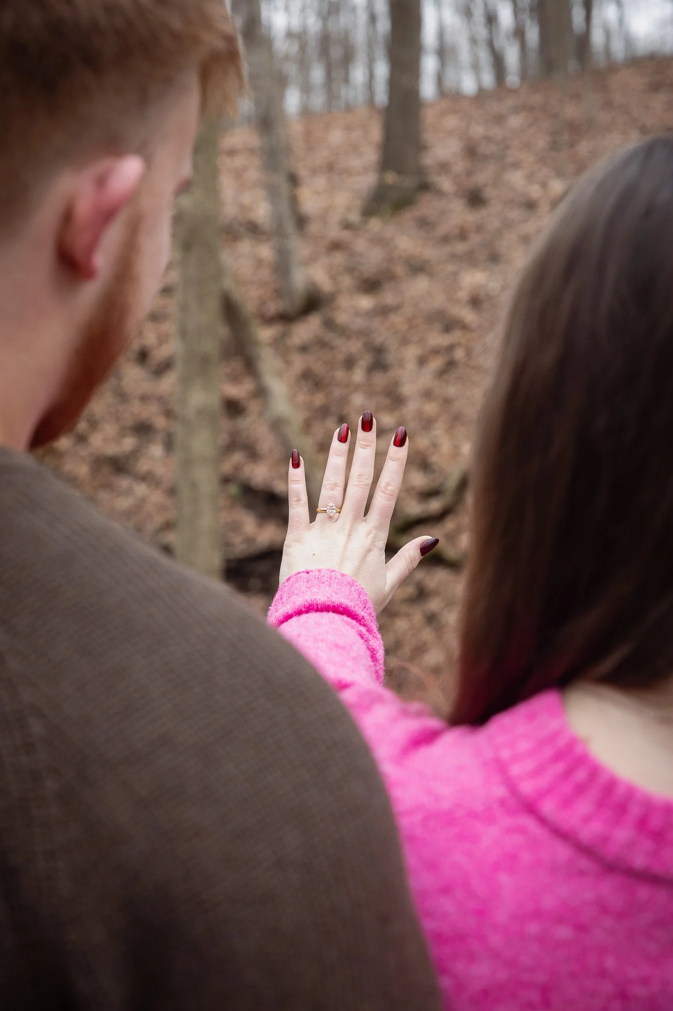 A man and woman outdoors in a wooded area, with the woman showcasing a ring on her ring finger, indicating an engagement or wedding.