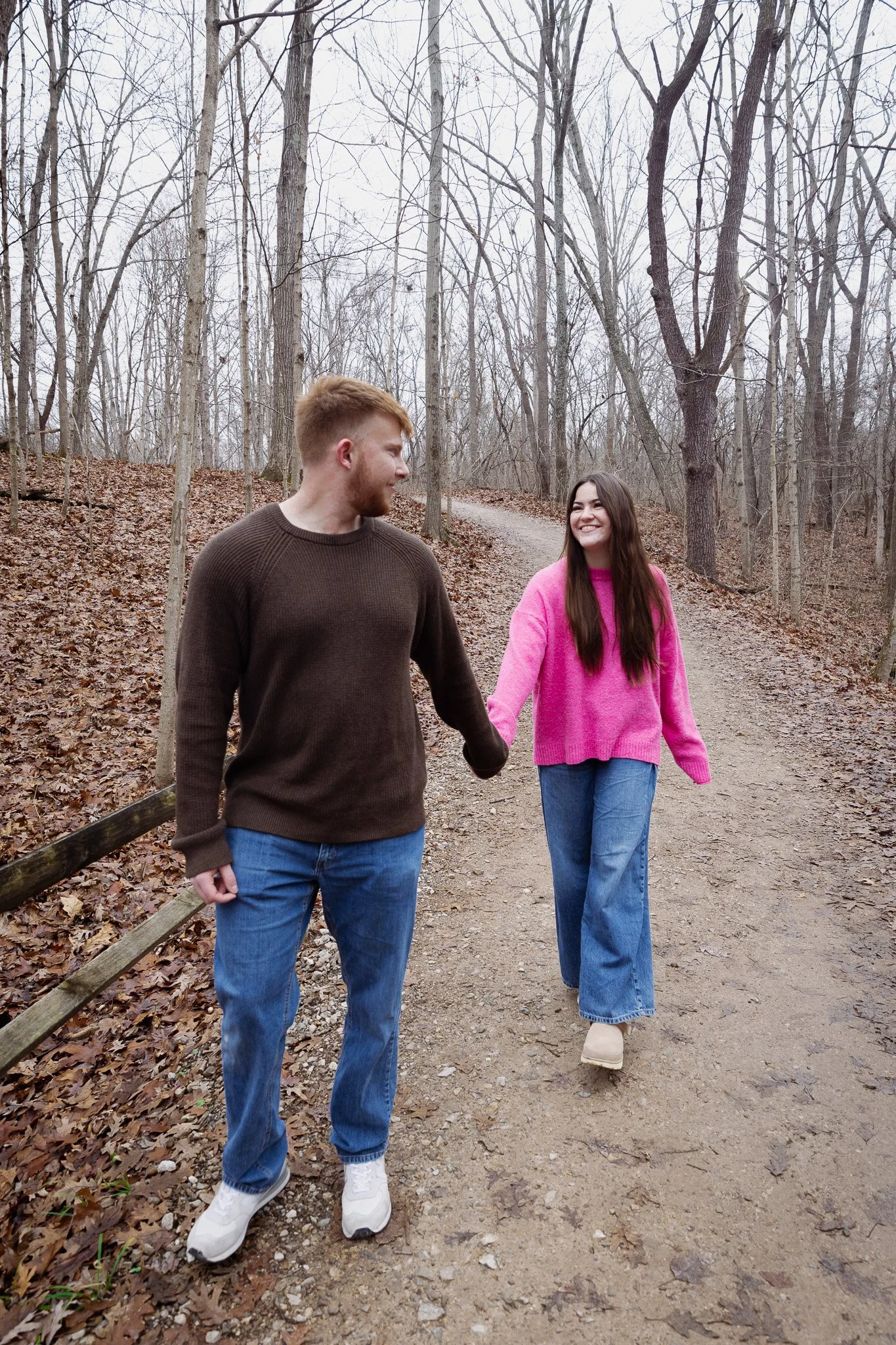 A young man and woman holding hands and walking on a dirt trail in a leafless forest, smiling at each other.