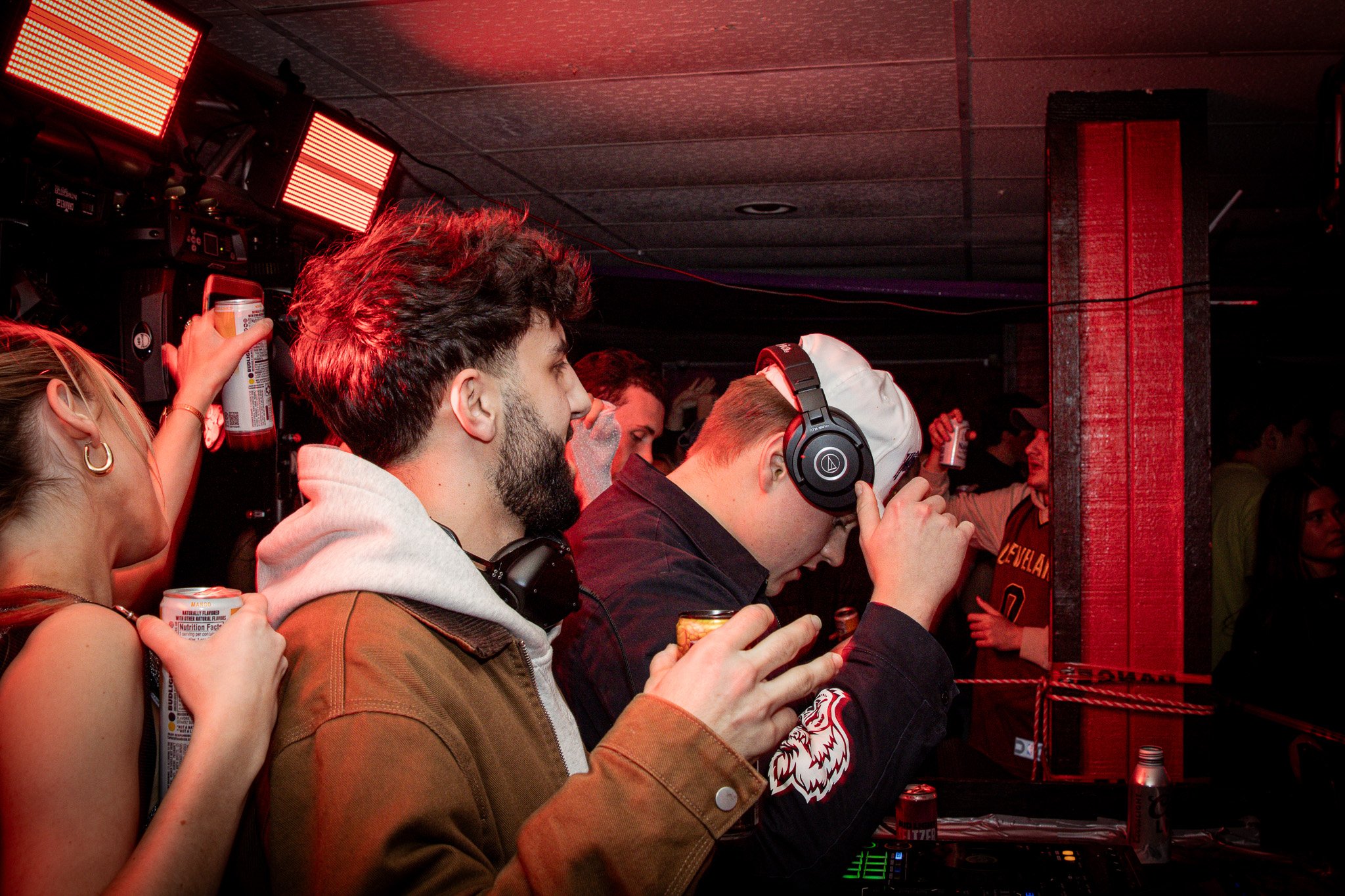 People at a crowded nightclub or music event, some holding drinks, with DJ equipment visible, illuminated by red lighting.