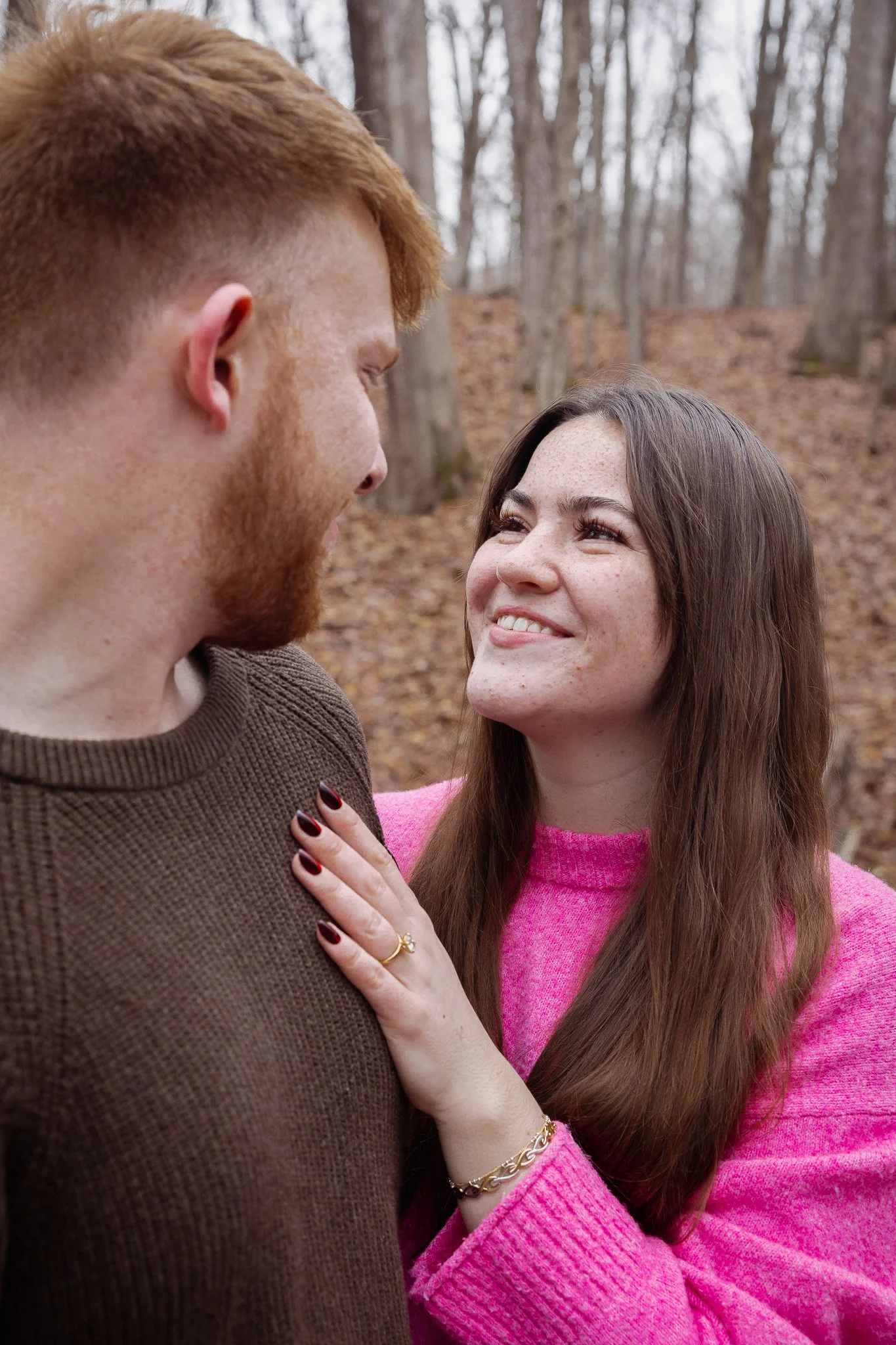 A man and woman smiling at each other outdoors in a wooded area with trees and fallen leaves. The woman has brown hair, freckles, and is wearing a pink sweater. The man has red hair and a beard, wearing a brown sweater.