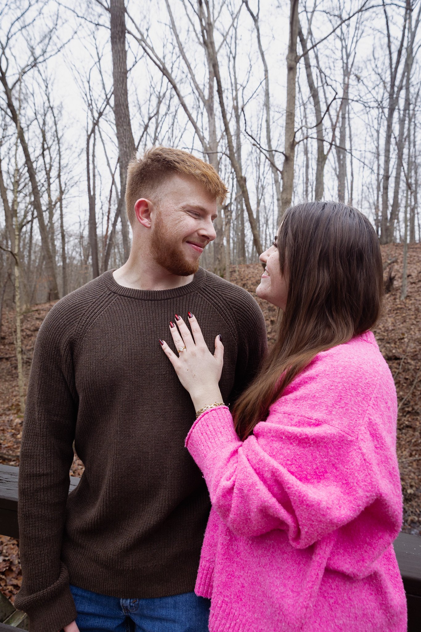 A man and woman sharing a happy moment outdoors in winter, with leafless trees in the background. The woman is touching the man's chest and they are smiling at each other.