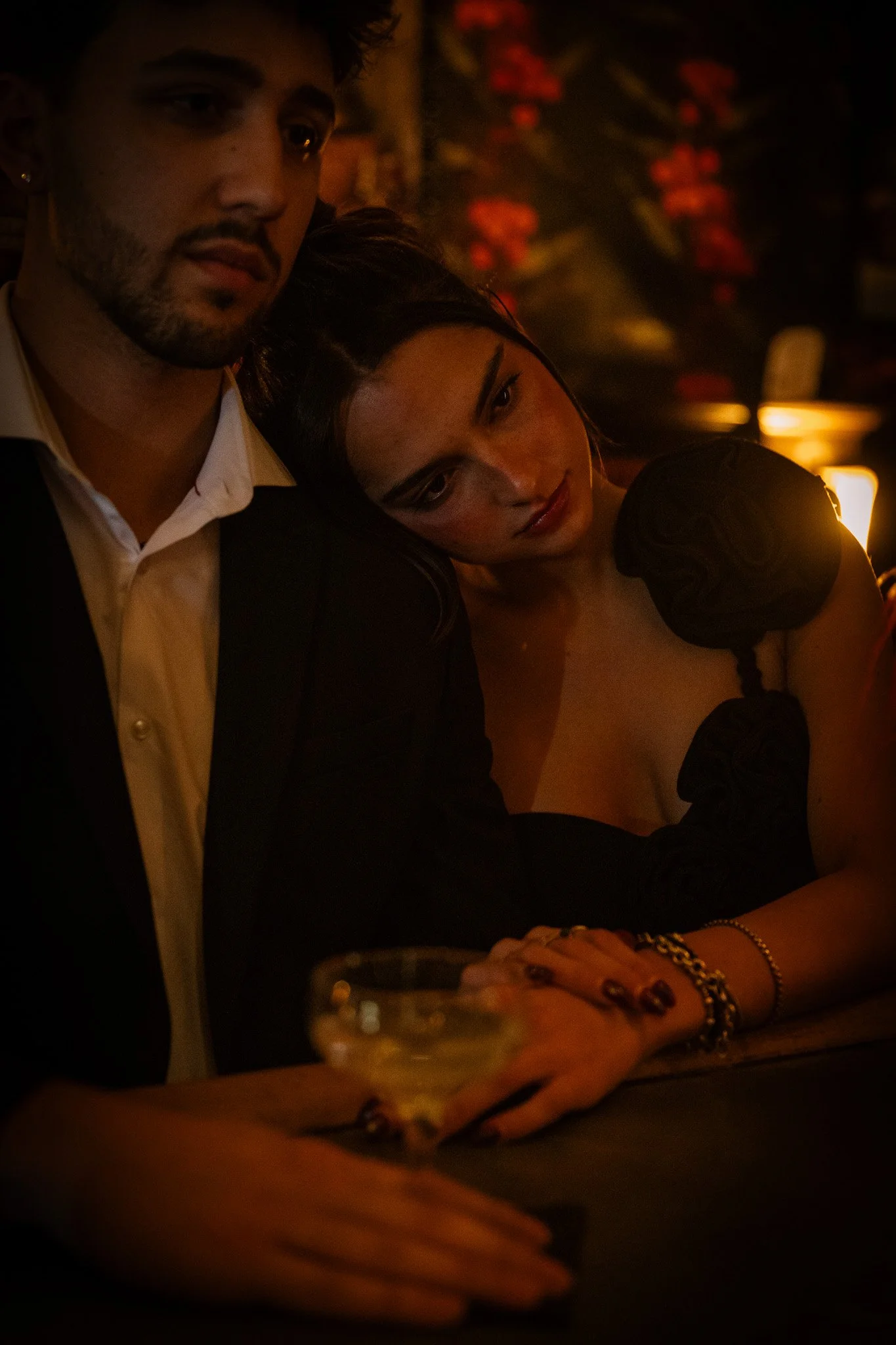 A man and a woman sit closely together in a dimly lit setting, with the woman's head resting on the man's shoulder, near a glass of orange drink on a dark table.