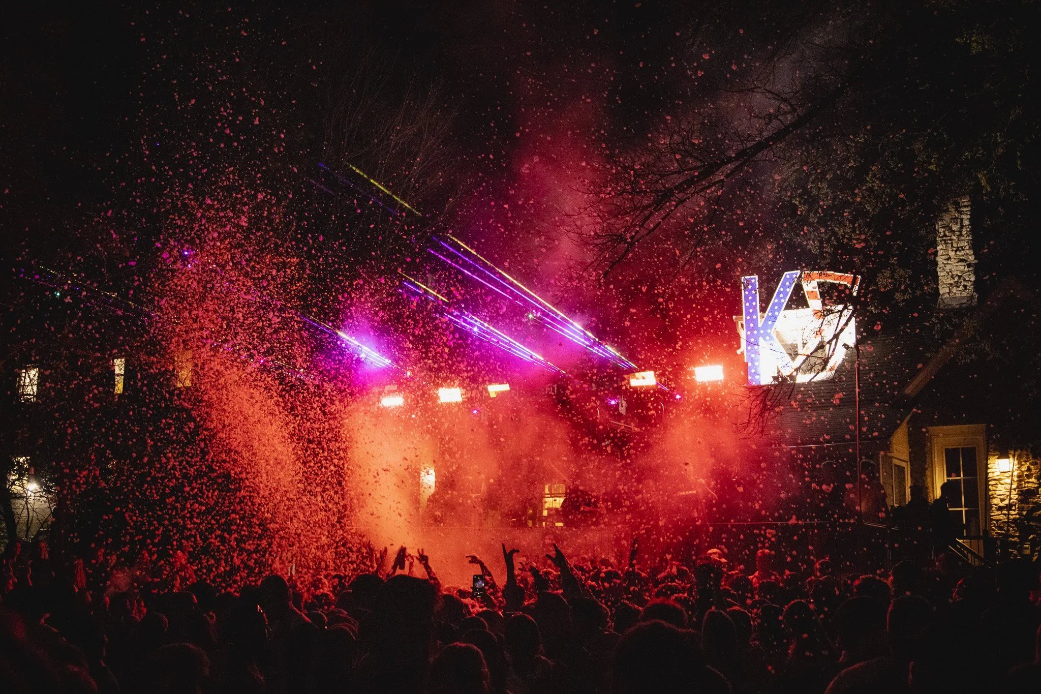 Crowd of people at a nighttime outdoor concert with confetti, colorful lights, and a large illuminated 'K' sign in the background.