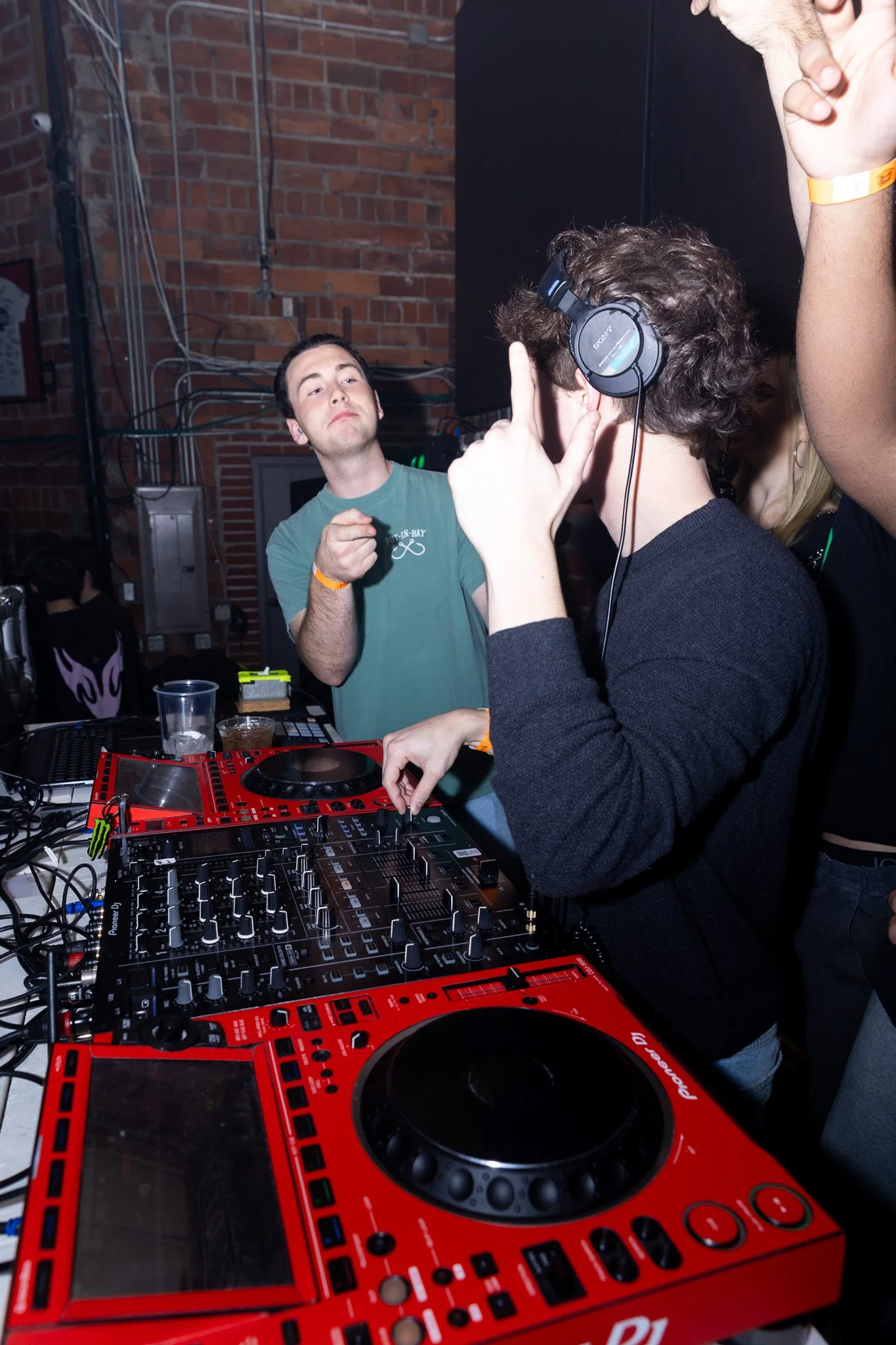A DJ with headphones adjusting music controls at a party, with another man pointing and making a gesture in front of brick wall and DJ equipment.