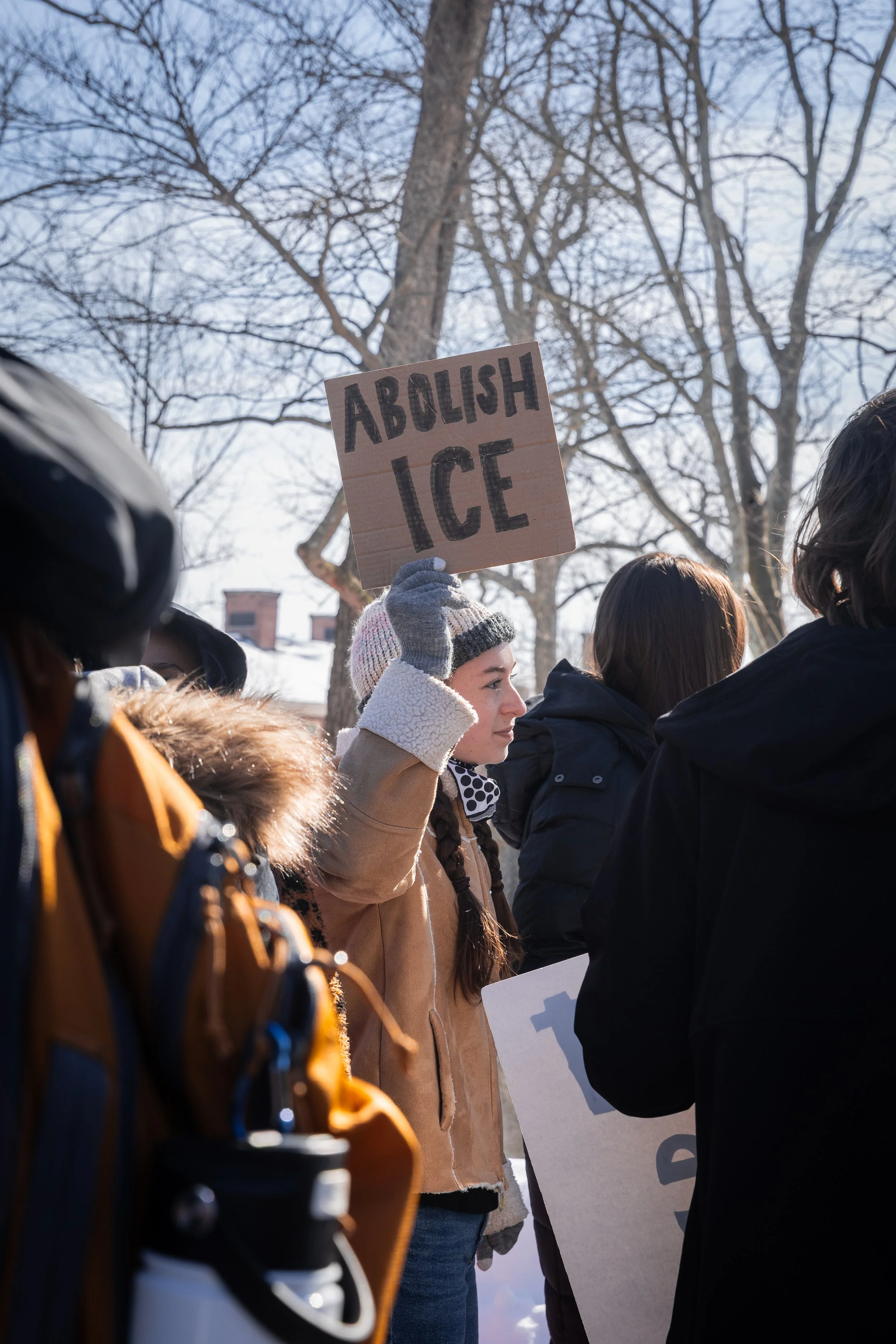 A young woman holds a sign that reads 'Abolish Ice' during an outdoor protest on a winter day.