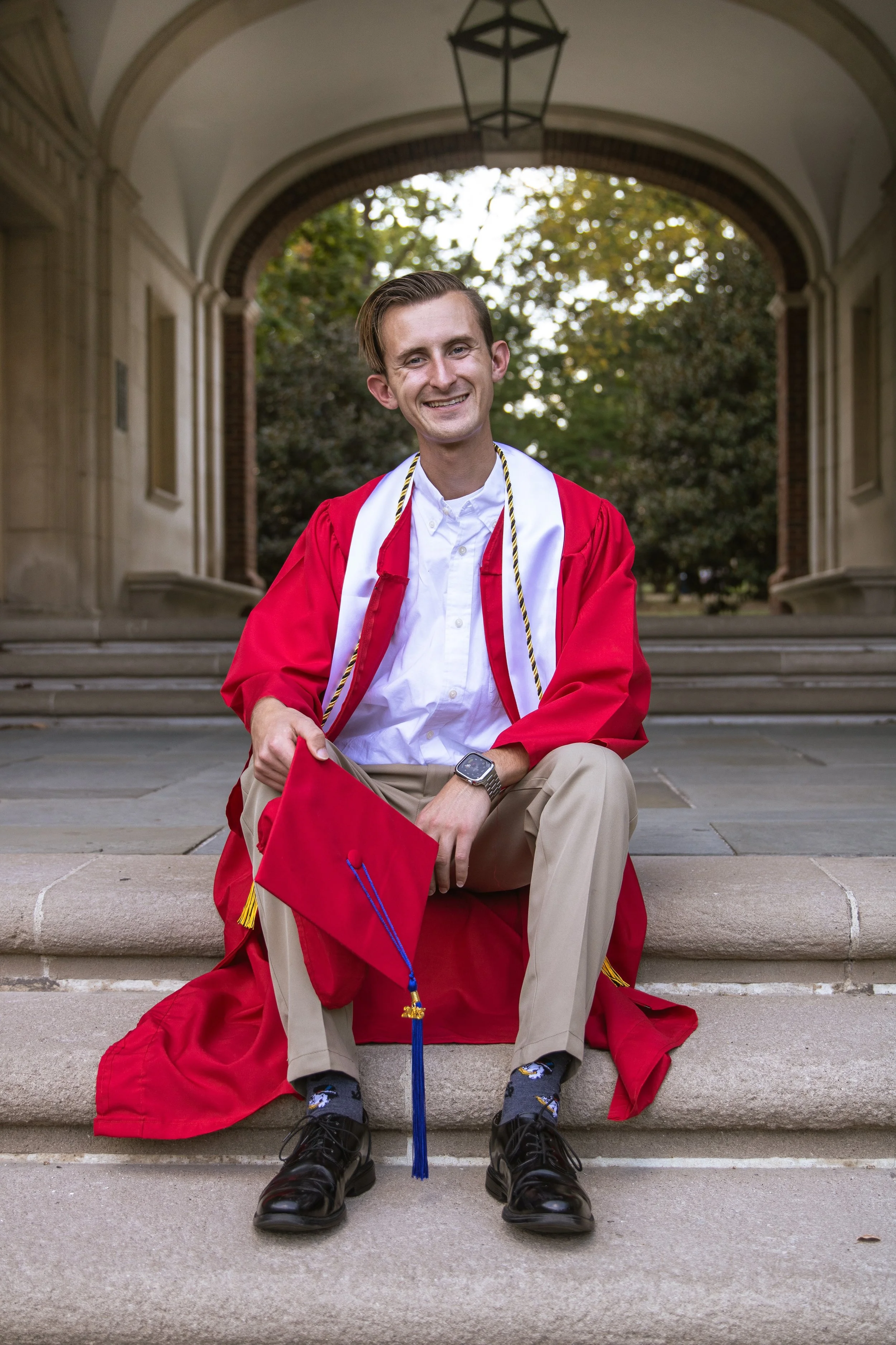 A young man in a red graduation gown and cap sitting on steps outside a building, smiling at the camera, holding his cap in his hand, with trees and an archway in the background.