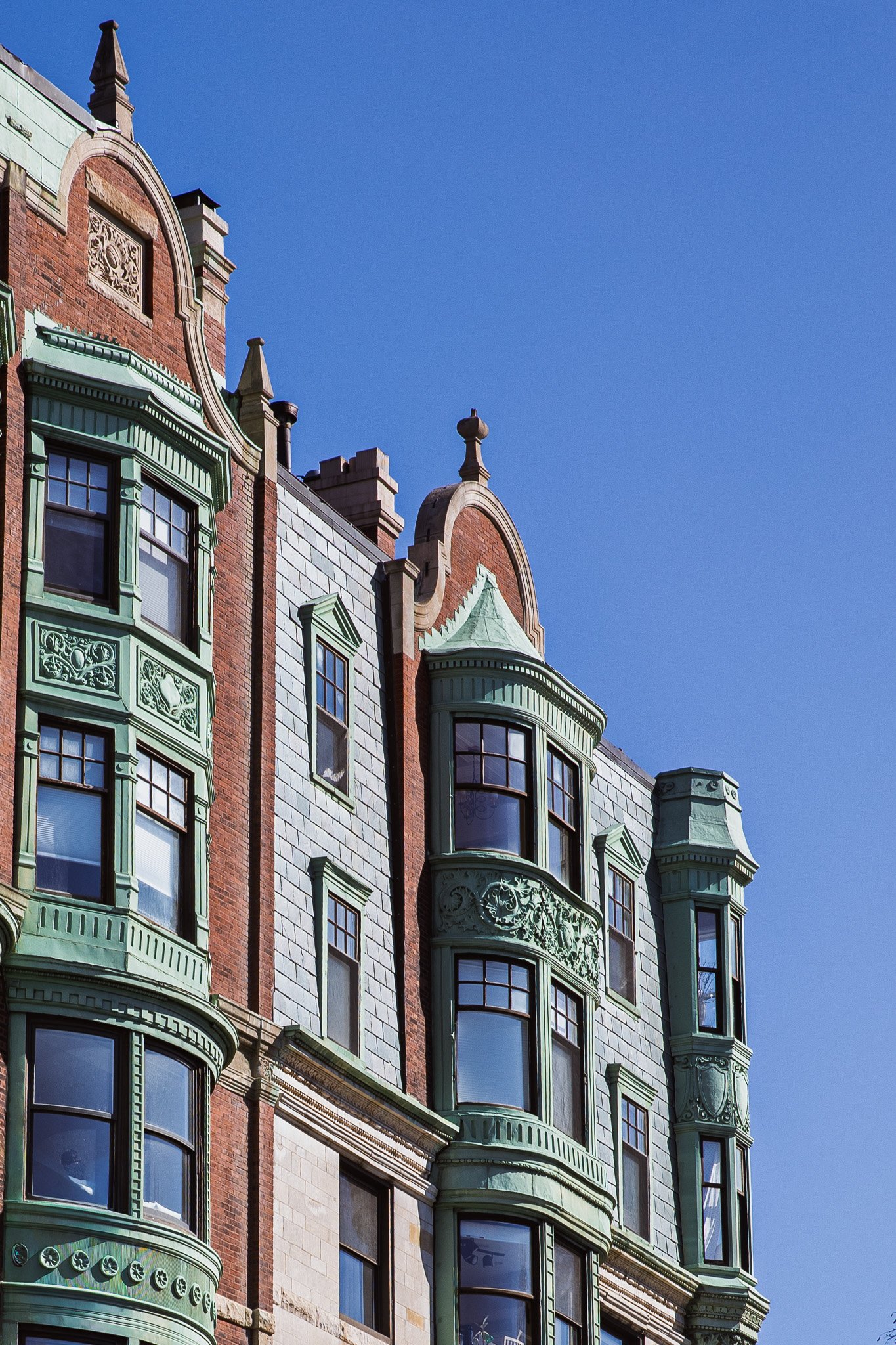 Close-up of an ornate old building with green decorative trim, bay windows, and brickwork, against a clear blue sky.