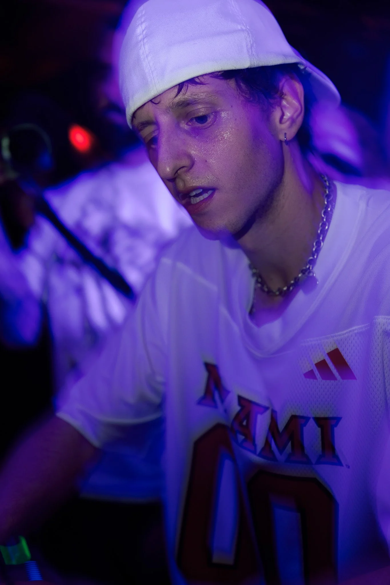 A young man with a white headscarf, wearing a white Miami sports jersey with red and blue lettering, appears to be at a party or club in a dimly lit environment with purple and blue lighting, sweating and focused.