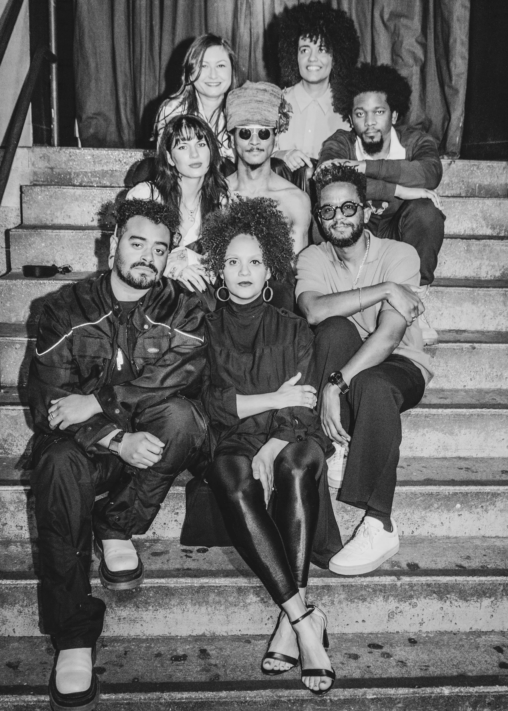 Black and white photo of a group of eight people sitting on a staircase, smiling. They appear to be casually dressed with a mix of modern styles.