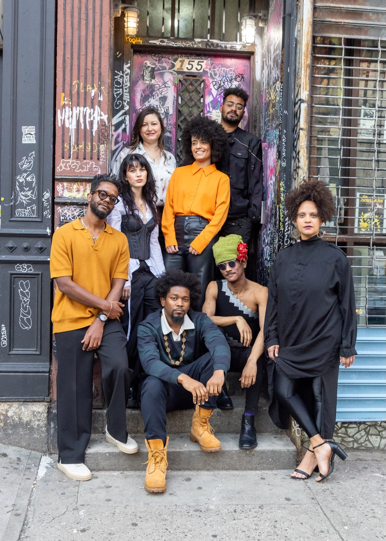 Group of eight diverse people posing in front of a graffiti-covered door, standing and sitting on steps, wearing stylish, eclectic clothing.