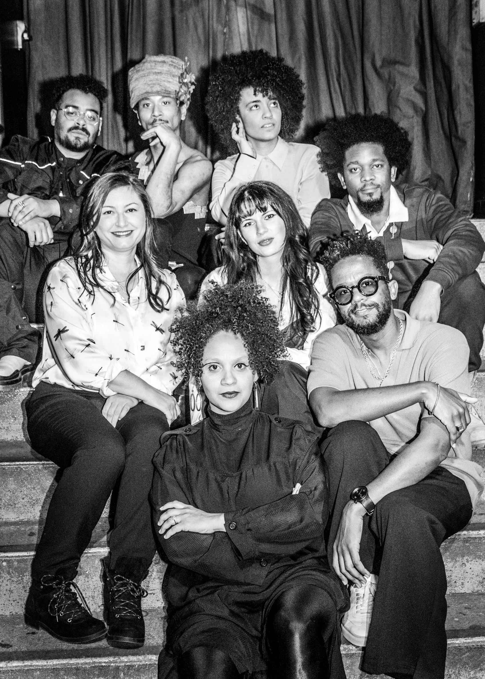 A diverse group of people sitting on stairs, posing for a black and white photo. They are dressed in varied styles, and some have curly hair. The setting appears casual and relaxed.