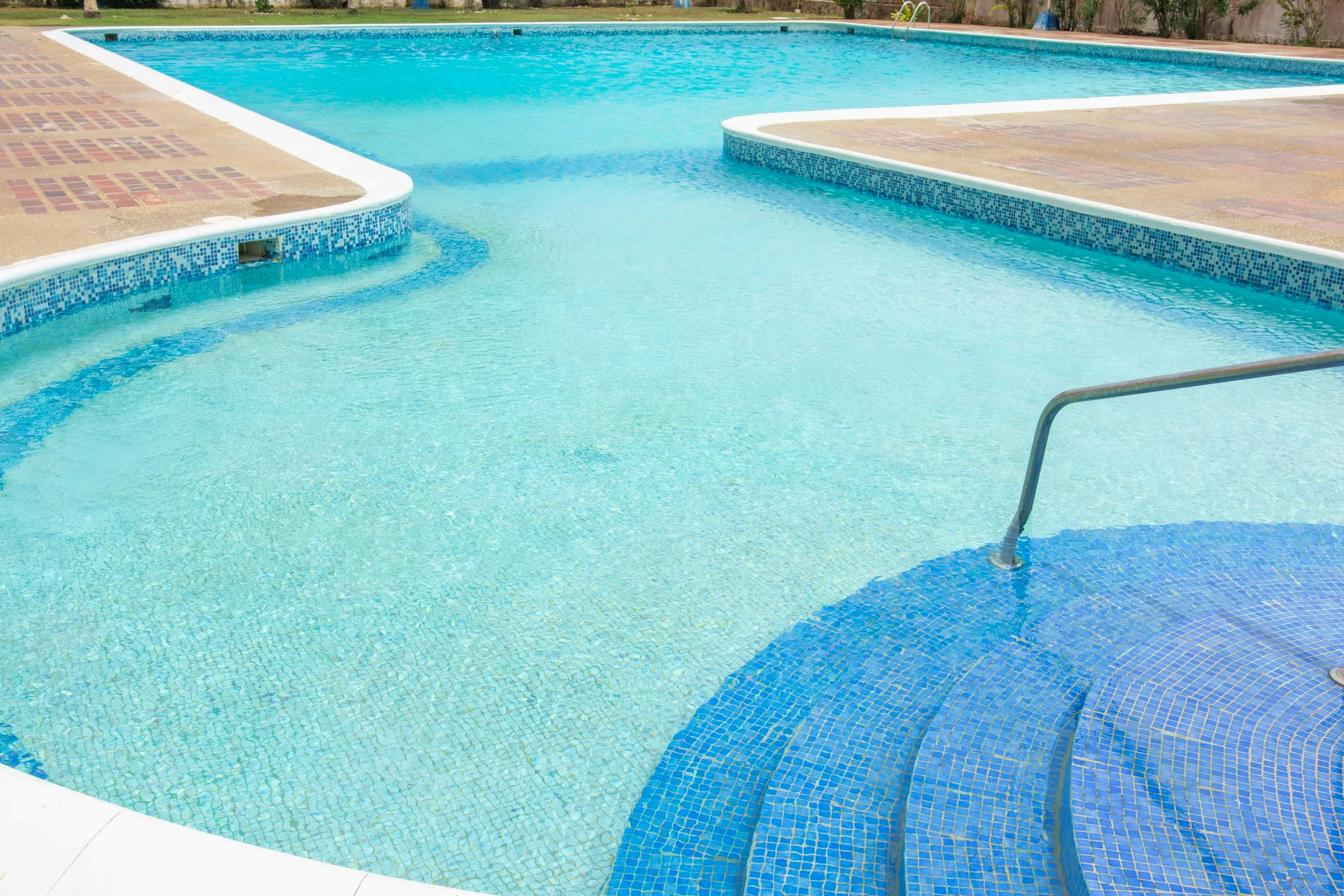 swimming pool with steps on the right and a ladder at the far end, surrounded by a paved deck and greenery.