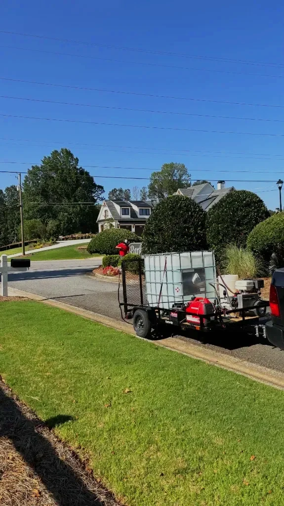 A trailer attached to a vehicle, with a large water storage tank and equipment, parked on a residential street with houses, trees, and a clear blue sky.