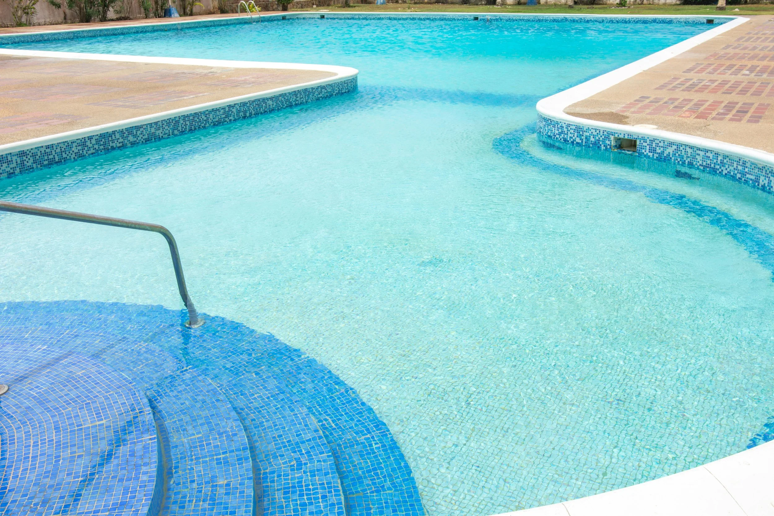 A swimming pool with clear blue water, surrounded by a concrete deck, with a set of ladder stairs on the left and a small submerged step area with blue mosaic tiles. The pool has a curving edge and is outdoors, with some grass and trees in the background.